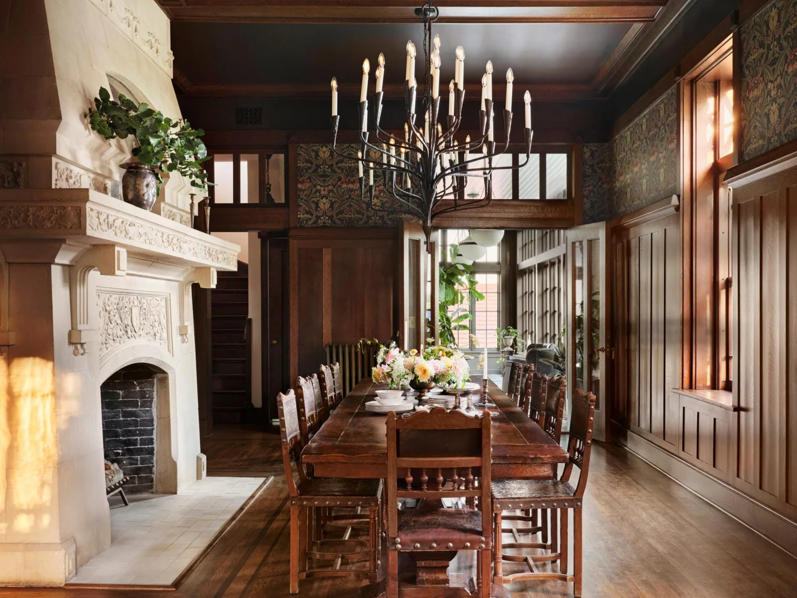 A formal dining room featuring a long wooden table, a large carved stone fireplace, dark wood wainscoting, and a multi-tiered black chandelier.