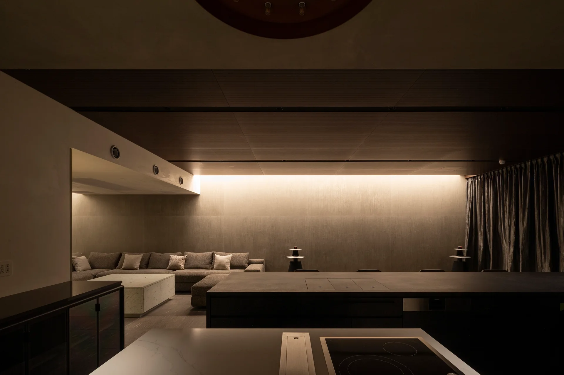 A moody open-plan living area features a dark wood paneled ceiling, a grey sectional sofa, and warm cove lighting casting a glow on a textured wall.