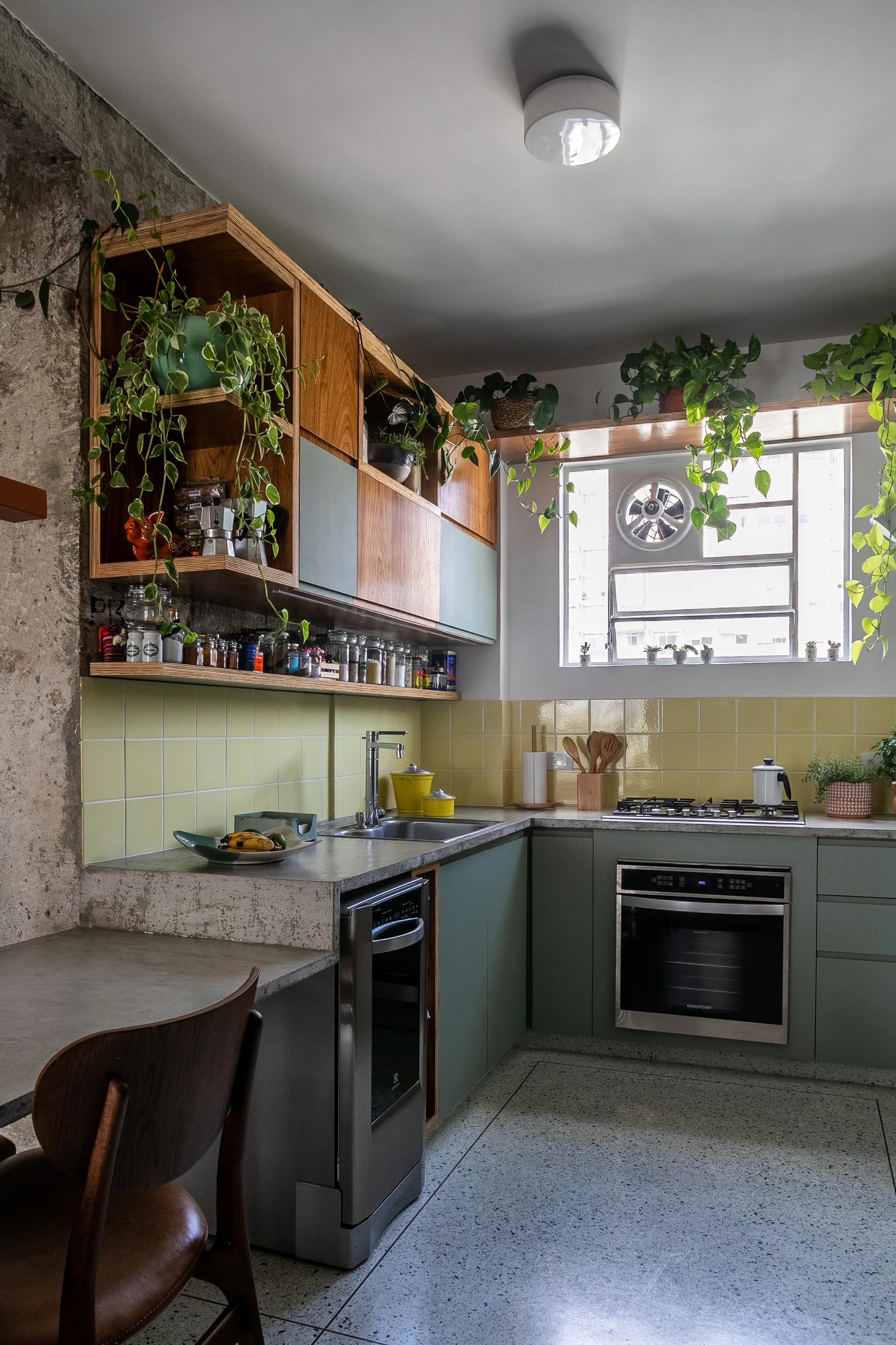 A kitchen features sage green cabinets, a yellow tiled backsplash, concrete countertops, and trailing plants on wood shelving.