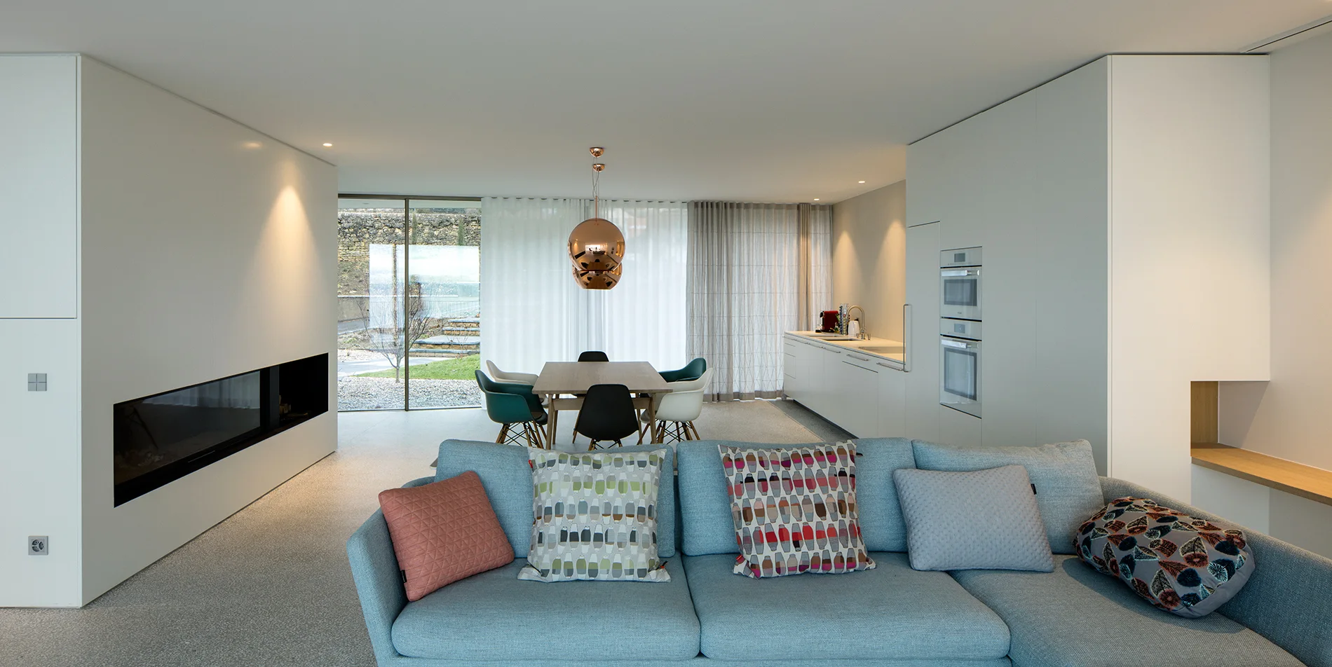 A modern open-concept living area features a light blue sofa with patterned pillows, a wood dining table, and built-in white kitchen cabinets.