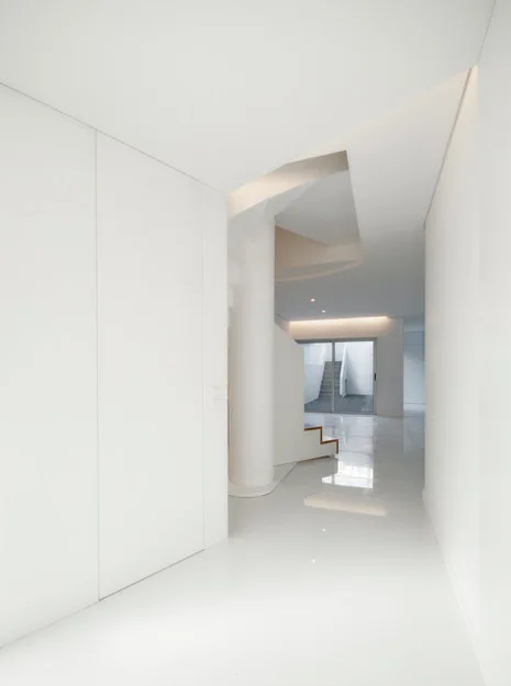 A minimalist white hallway featuring a highly reflective polished floor, a central cylindrical column, and a modern staircase in the background.