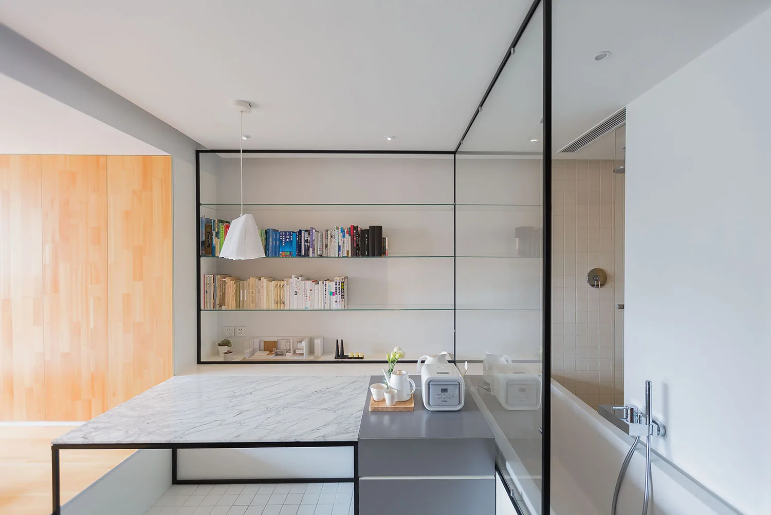 A modern bathroom features a glass partition separating a bathtub from a marble-topped vanity and black-framed open shelves filled with books.
