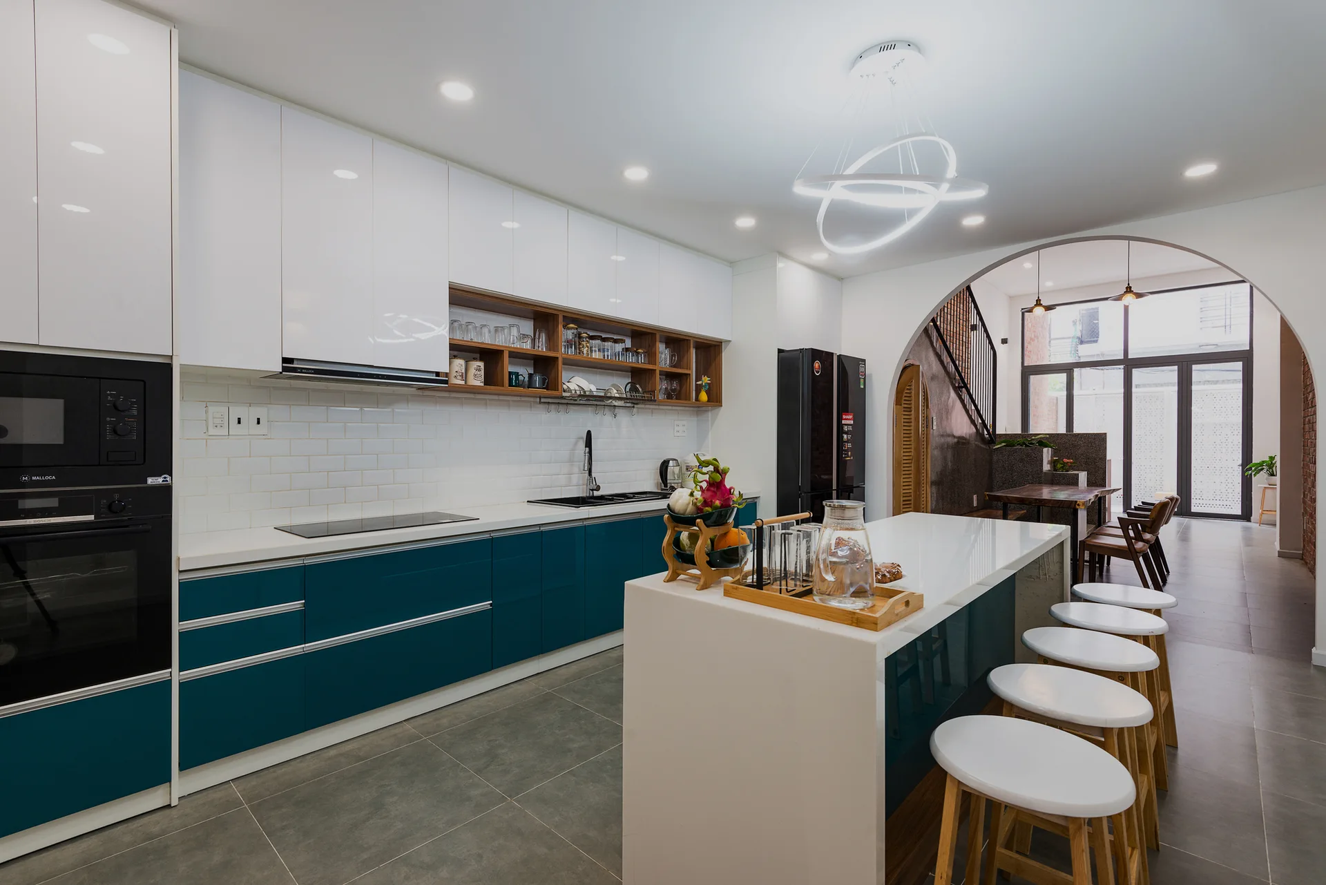 A modern kitchen features glossy teal base cabinets, white upper cabinetry, a central white island with stools, and an open archway leading to another room.