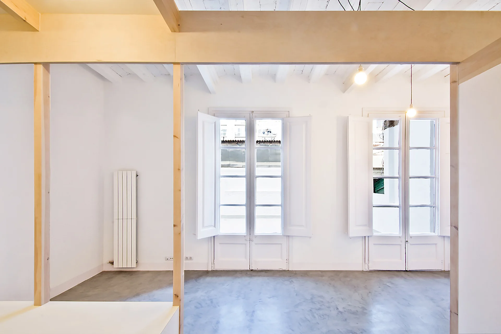 An empty minimalist room with white walls, concrete floors, exposed light wood timber framing, and tall windows with matching white folding shutters.