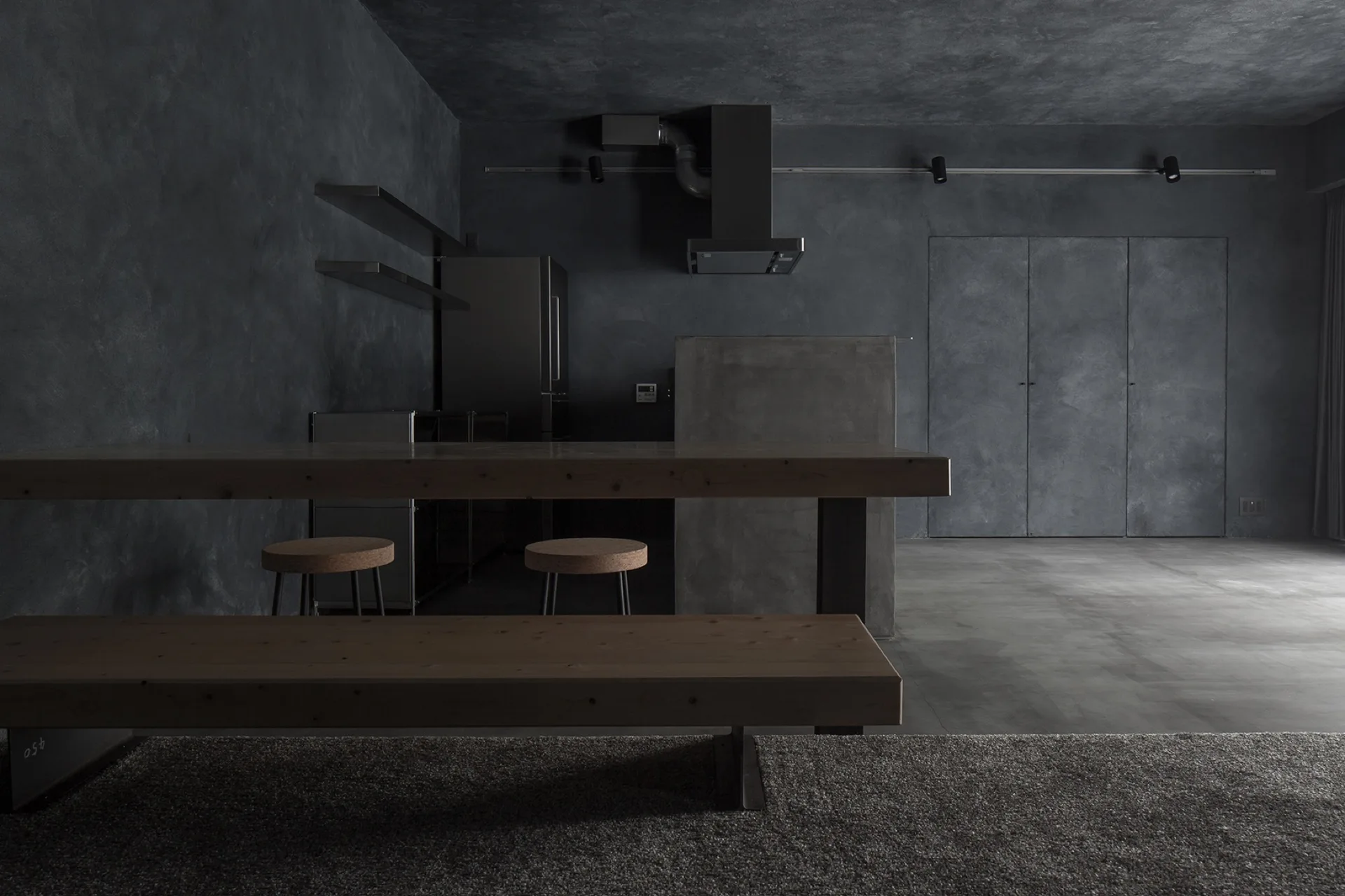 A dark minimalist kitchen features charcoal textured plaster walls, a concrete island, and a long wood dining table with a matching bench.