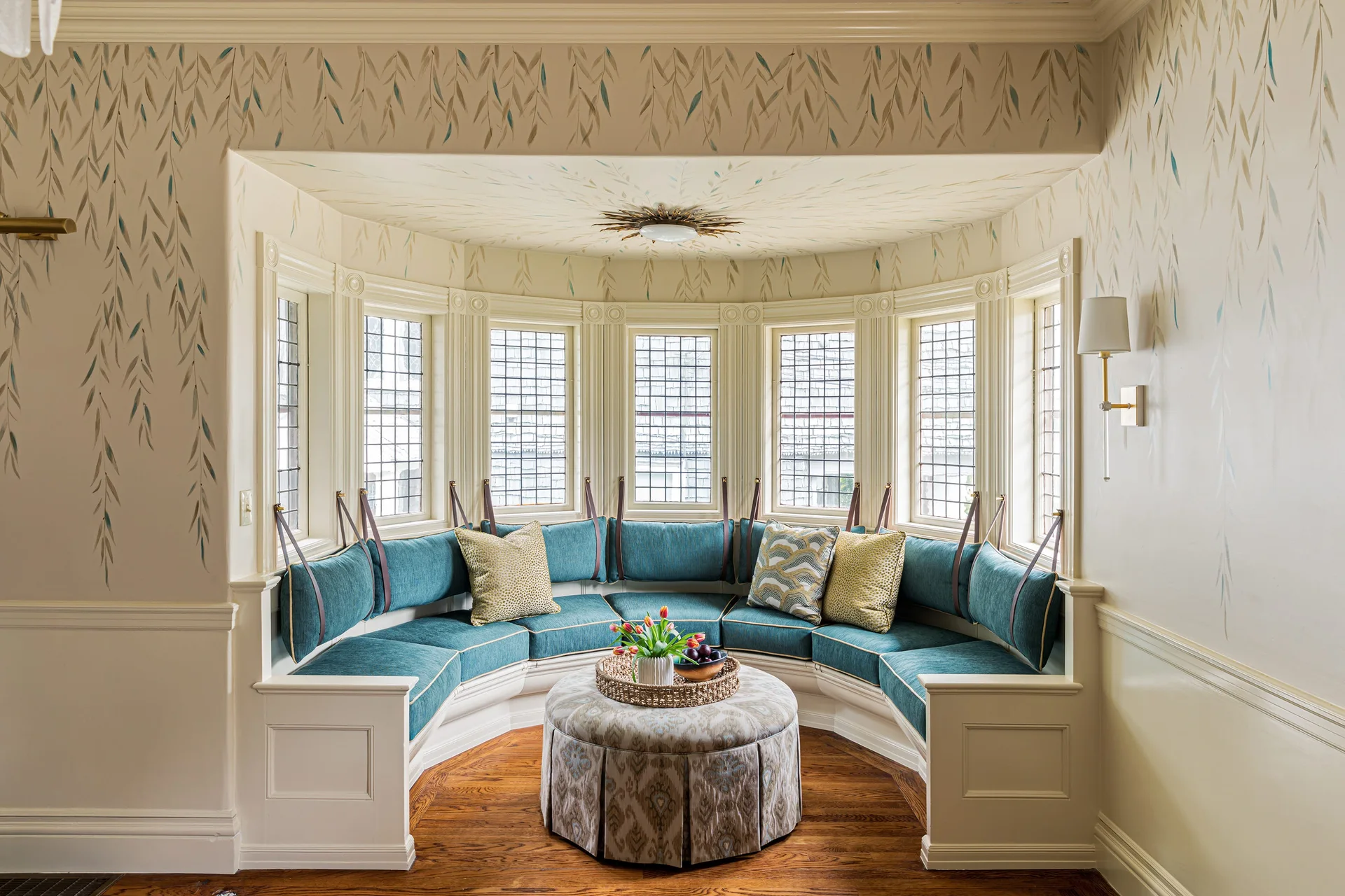 A curved built-in bench with teal cushions sits in a bay window alcove under cream-colored willow branch wallpaper with an upholstered center ottoman.