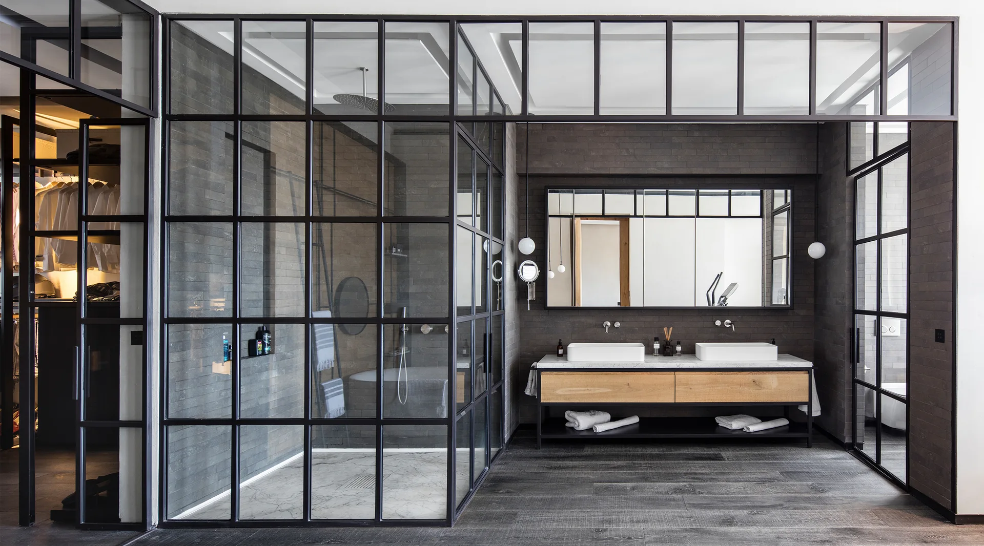 A modern industrial bathroom features a black steel grid glass shower enclosure, a wood double vanity, and dark grey tiled walls.