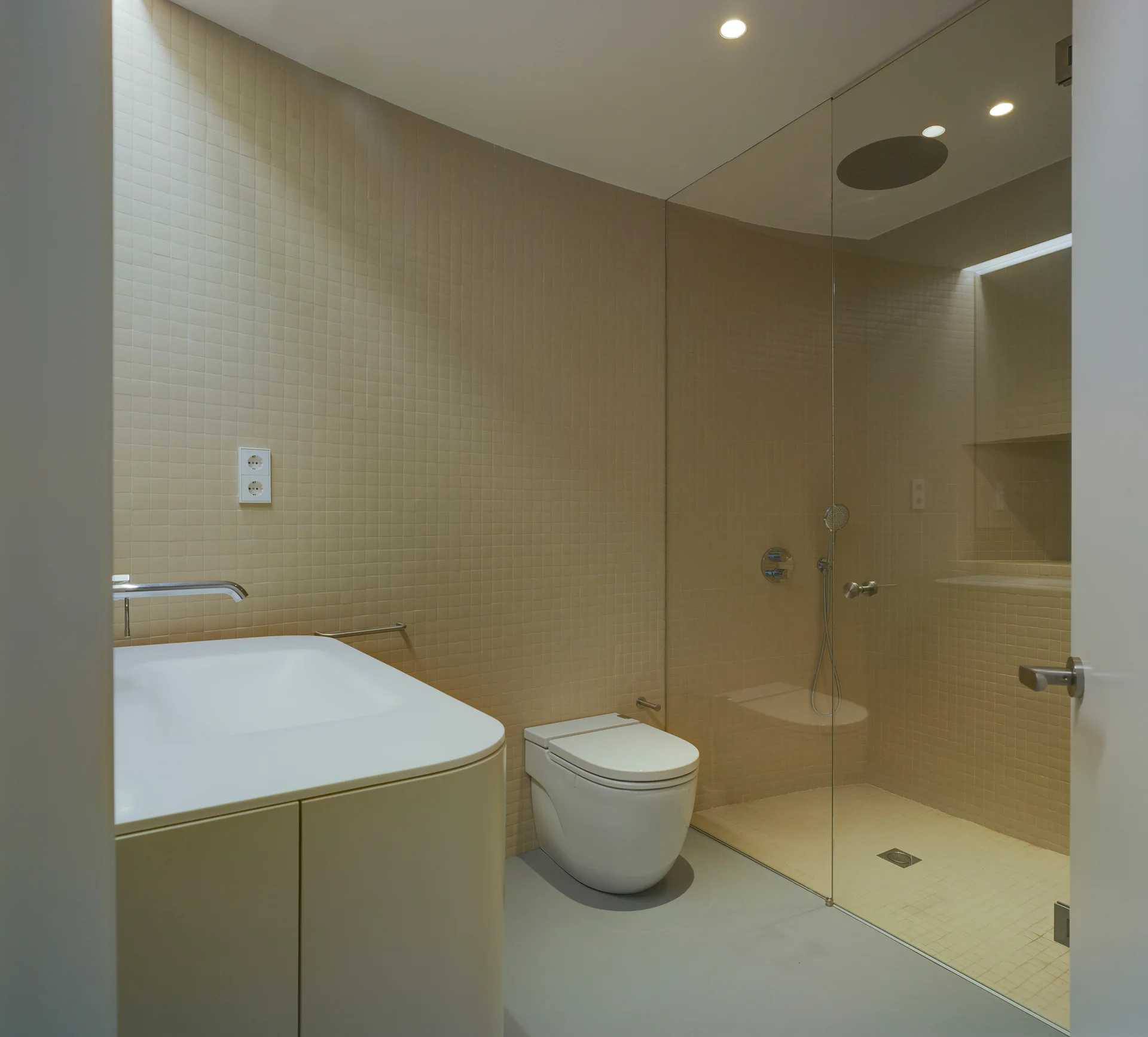 A minimalist bathroom features beige square-tiled walls, a curved white vanity, a wall-mounted toilet, and a glass-enclosed walk-in shower.