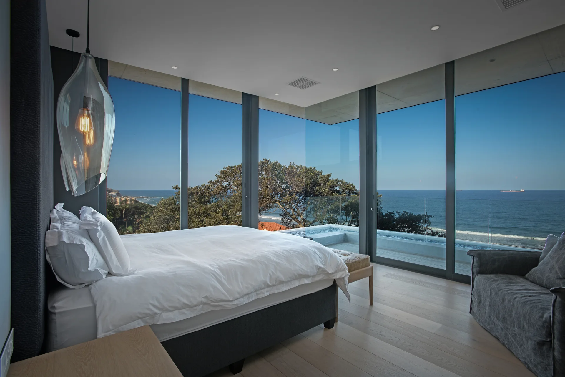 A minimalist bedroom features a bed with white bedding and a gray armchair next to floor-to-ceiling glass windows with an ocean view.