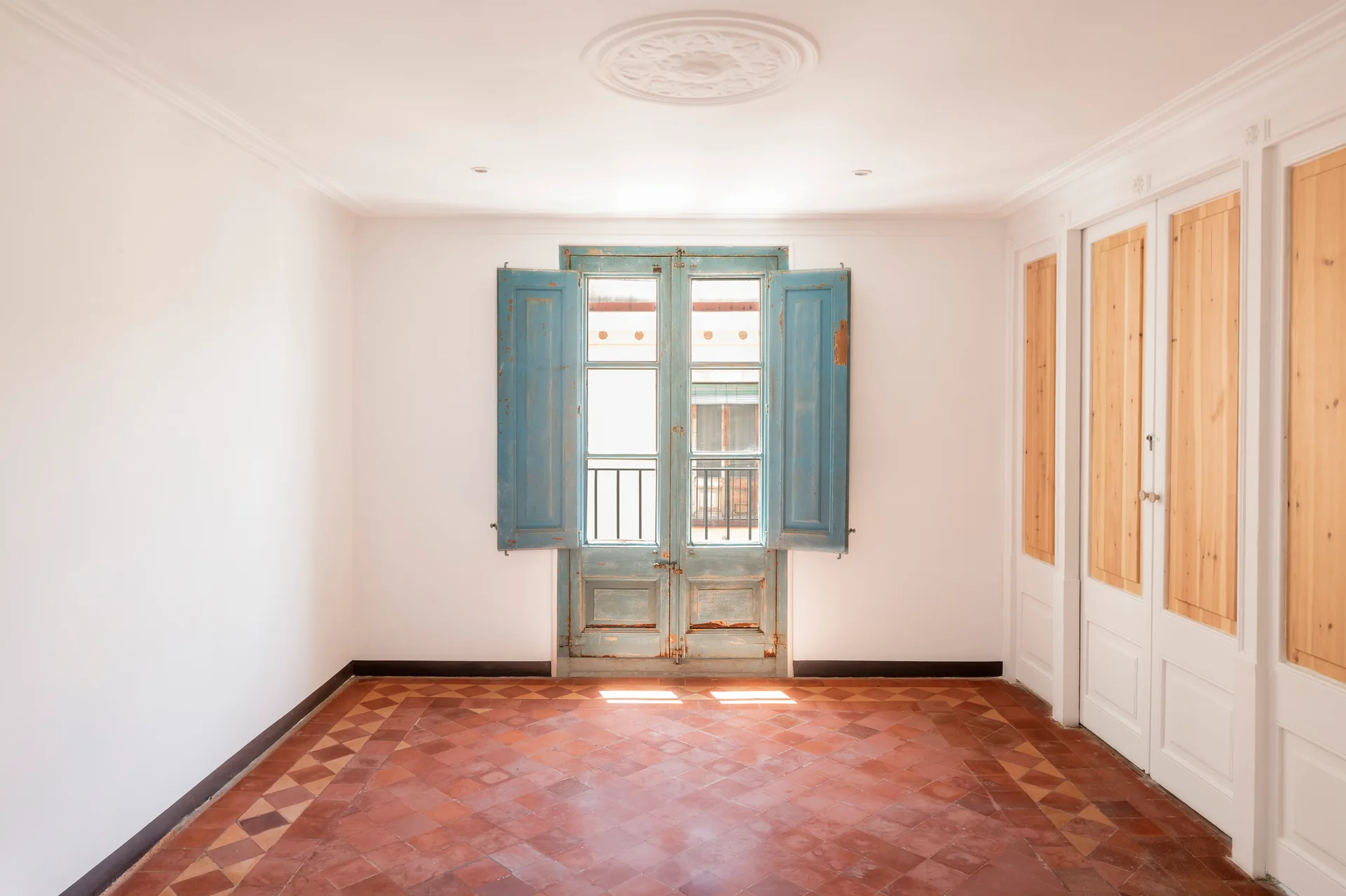 An empty room featuring terracotta tile flooring, white walls, built-in wooden cabinets, and a central double door with distressed blue shutters.