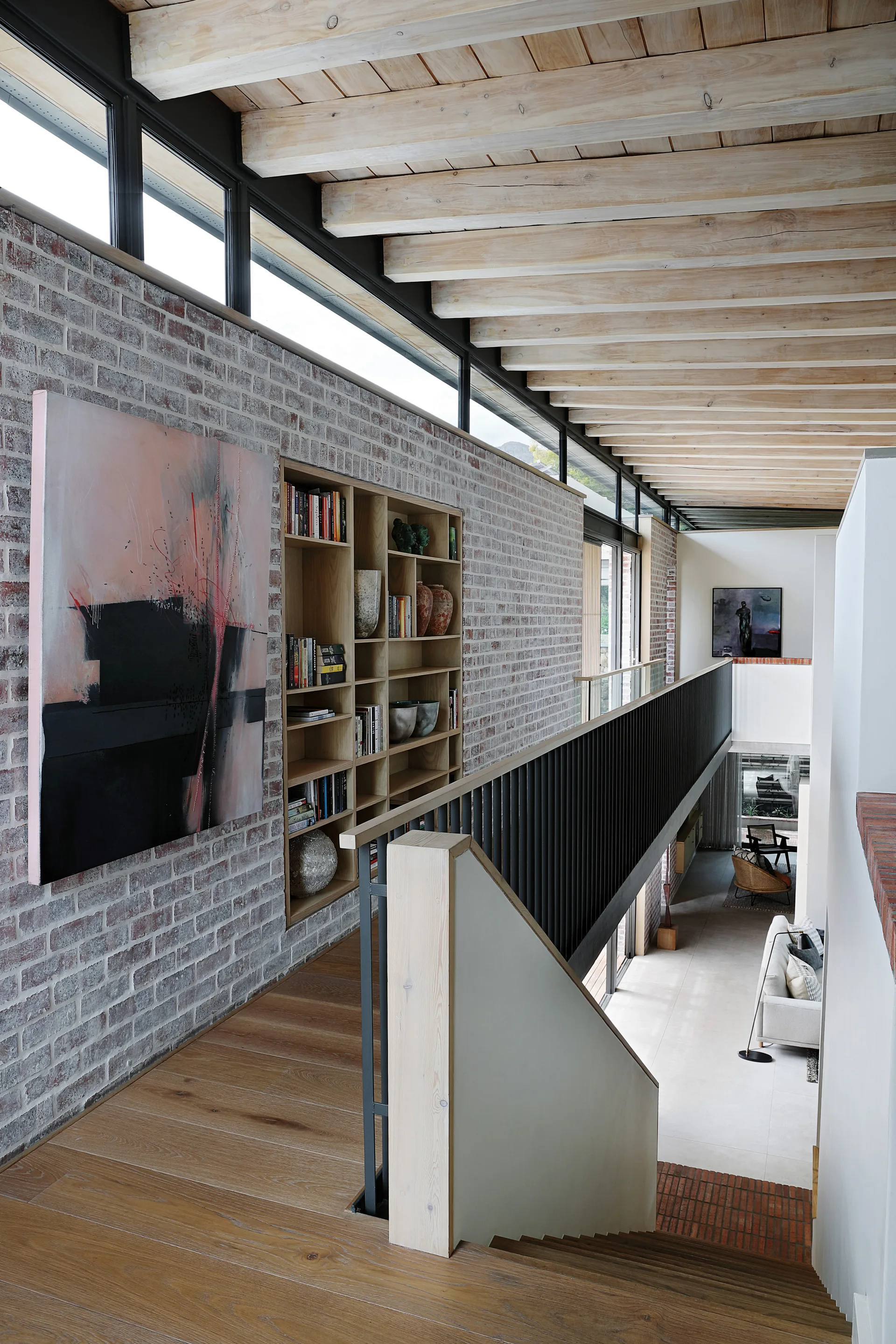 A modern mezzanine landing features a whitewashed brick wall with built-in wooden bookshelves, exposed ceiling beams, and a black metal railing.