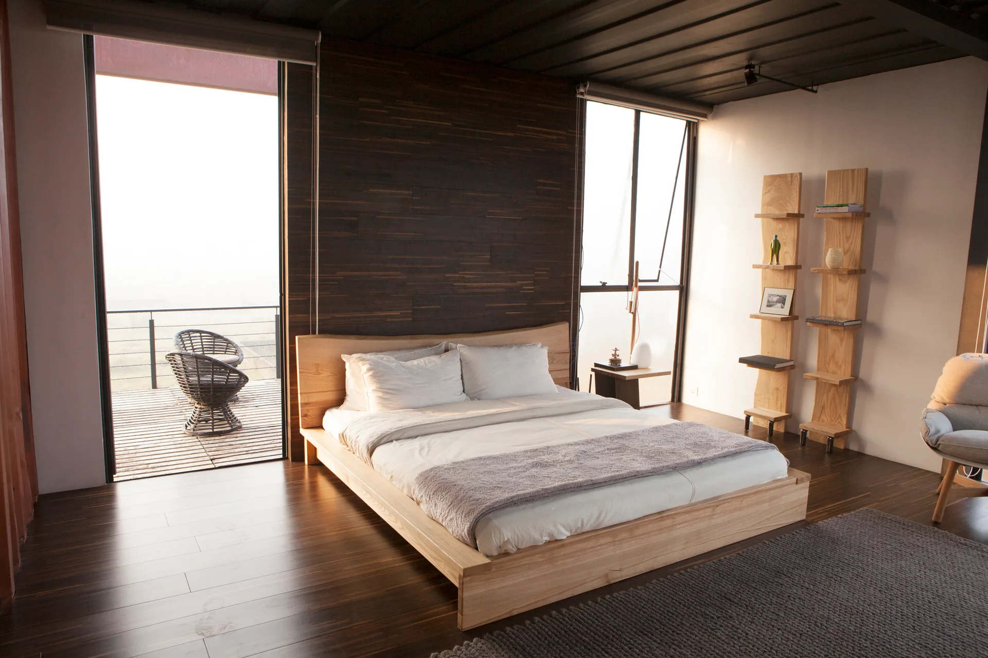 A modern bedroom features a light wood platform bed, a dark wood-paneled feature wall, and tall windows next to a private balcony.
