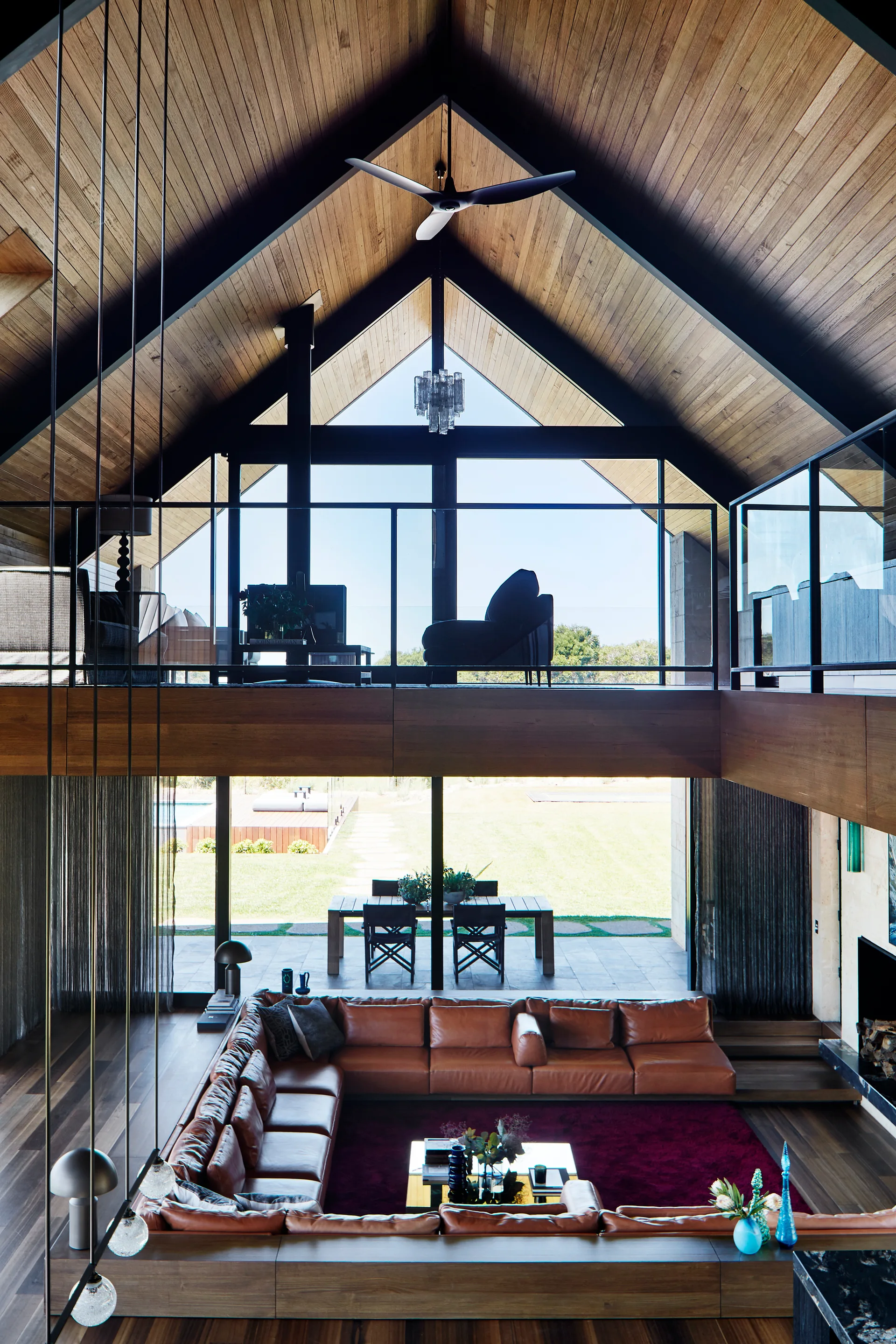 A high-angle view of a sunken living room featuring a U-shaped brown leather sectional, a red rug, and a vaulted wood plank ceiling.
