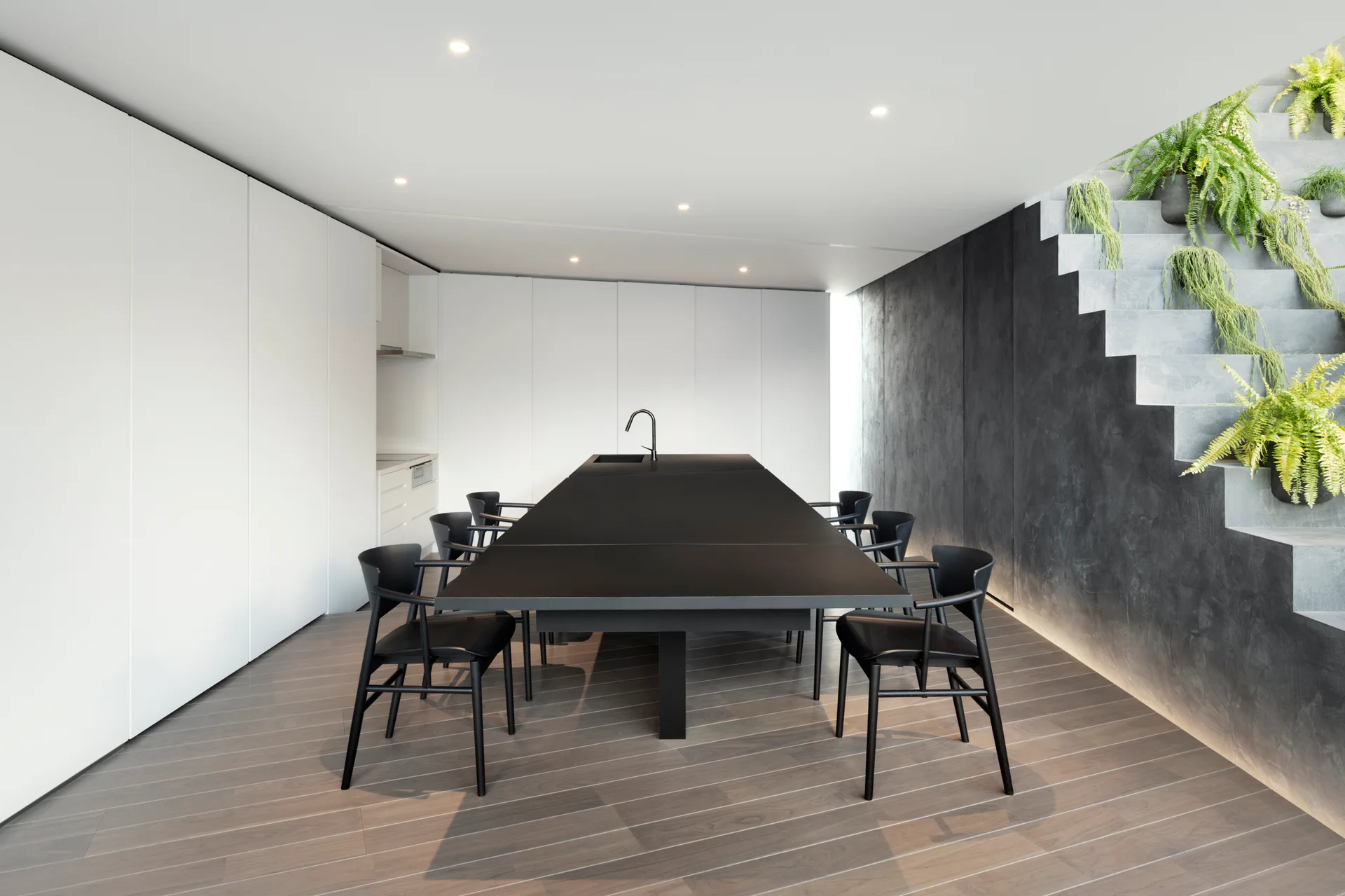 A long black kitchen island and chairs sit in a minimalist kitchen with white cabinets and a concrete staircase decorated with potted green plants.