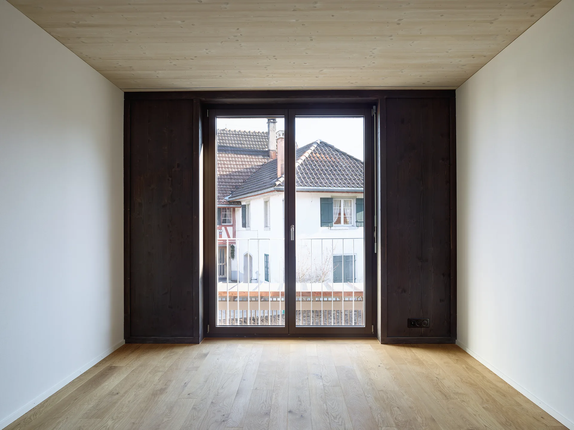 An empty room features light wood flooring, a matching wooden ceiling, white walls, and a large window flanked by dark wood panels.