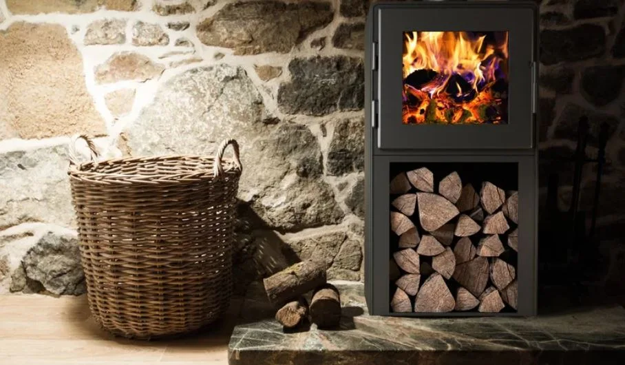 A black metal wood burning stove with integrated log storage sits on a dark stone hearth against an irregular stone masonry wall next to a wicker basket.