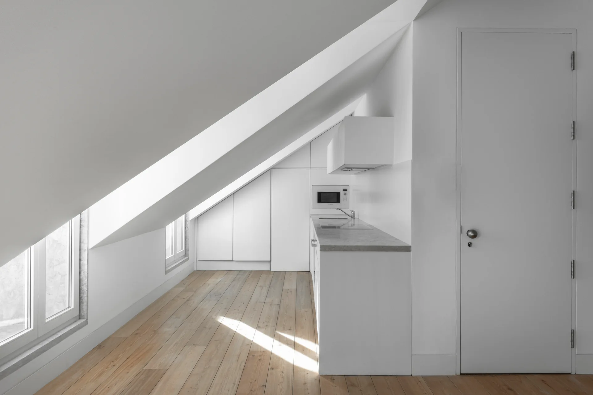 A minimalist attic kitchen features white handleless cabinetry fitted beneath a sloped ceiling with light wood plank flooring and a gray countertop.