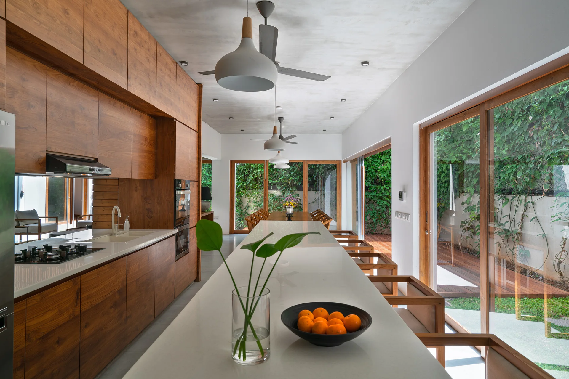 A modern kitchen featuring warm wood-grain cabinets, a long white stone-topped island, and sliding glass doors that open to a lush garden.