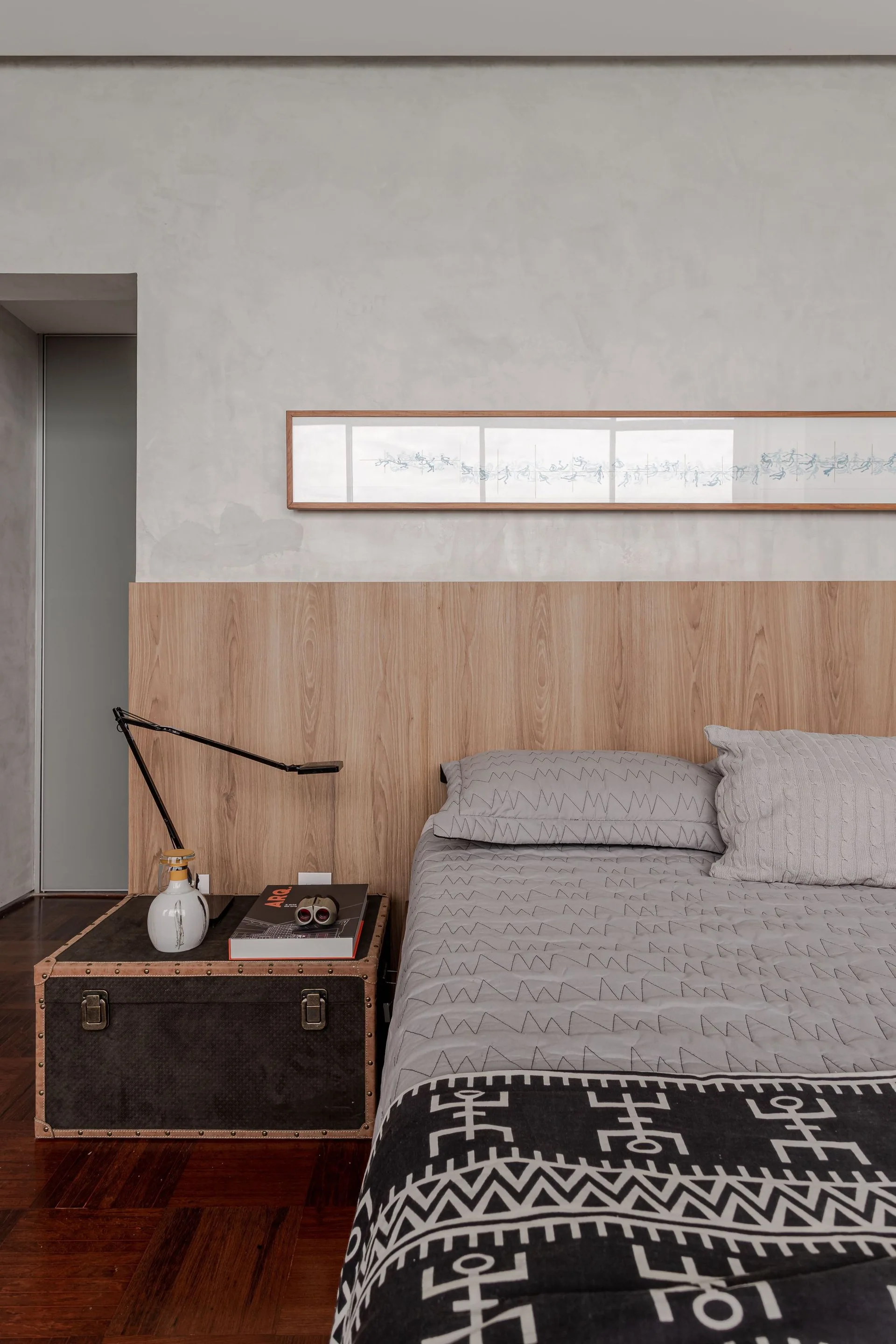 A modern bedroom features gray quilted bedding, a half-wall wood headboard panel, and a vintage-style trunk used as a bedside table.
