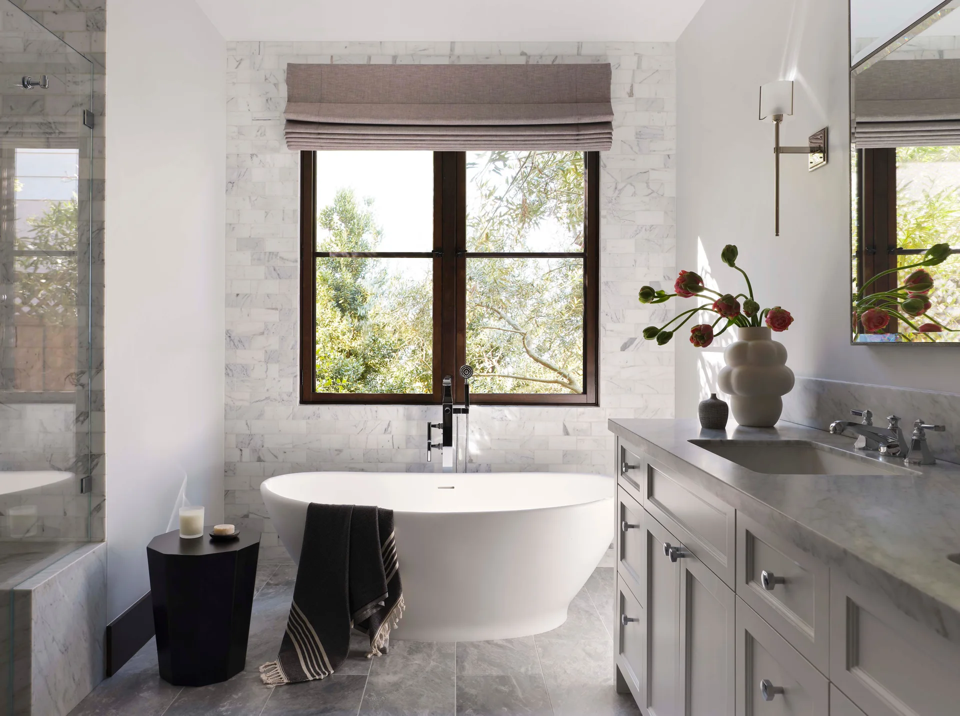 A white freestanding bathtub sits under a window framed by marble subway tiles, next to a grey double-sink vanity with marble counters.