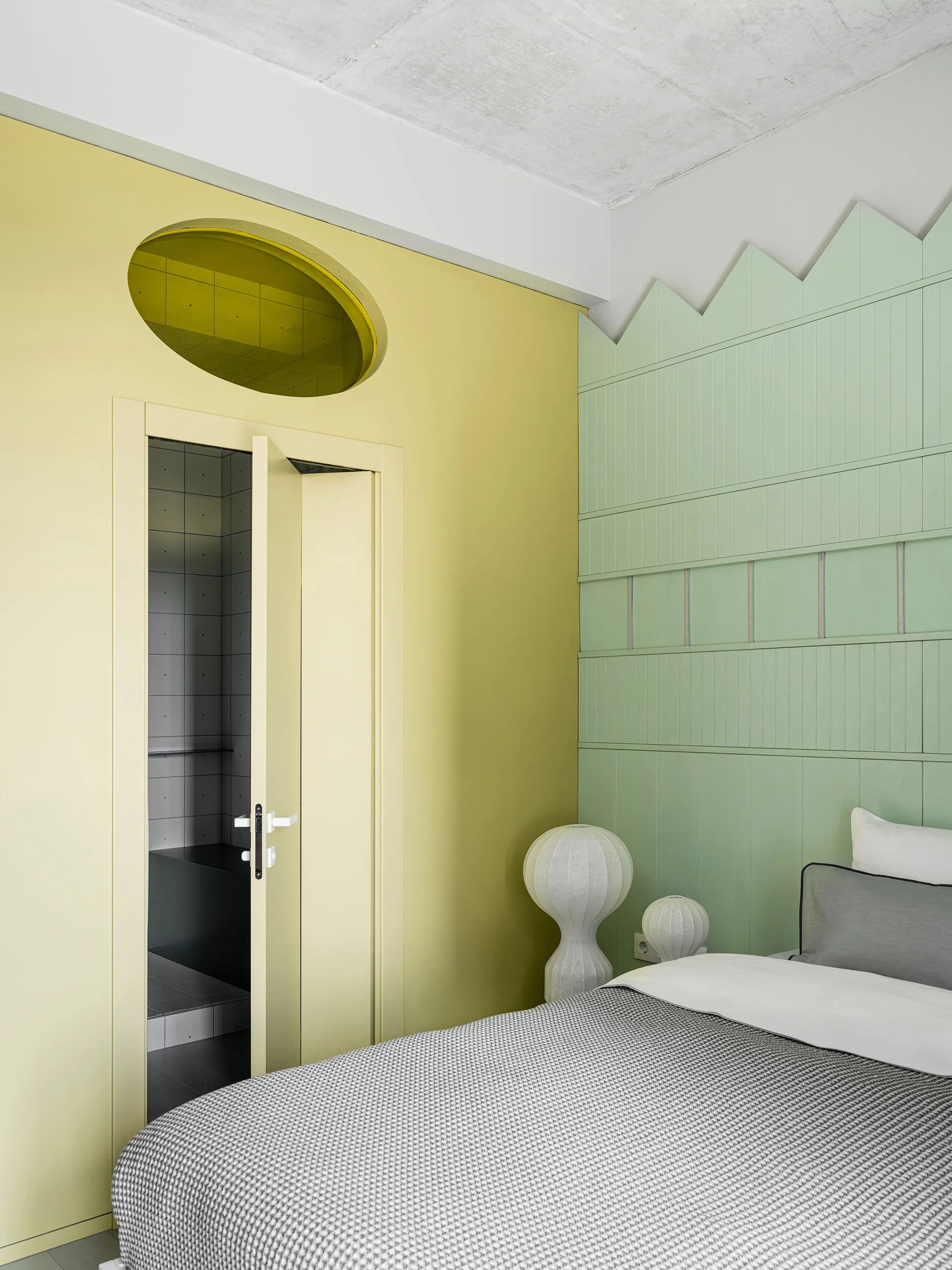 A modern bedroom features a grey bed set against a sage green paneled wall and a yellow accent wall with an oval transom window above the door.