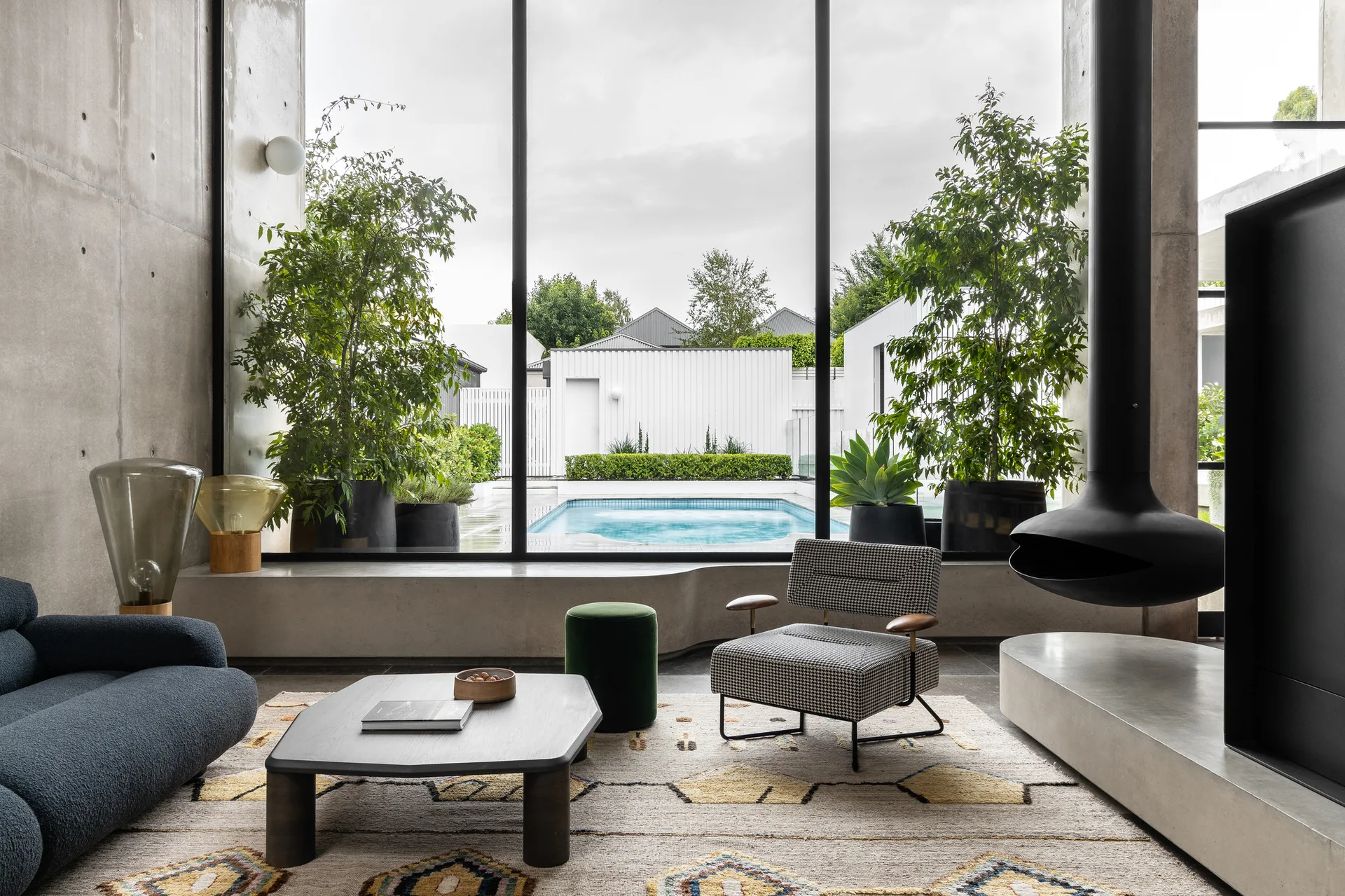 A modern living room features raw concrete walls, a large window overlooking a backyard pool, a suspended black fireplace, and a patterned armchair.