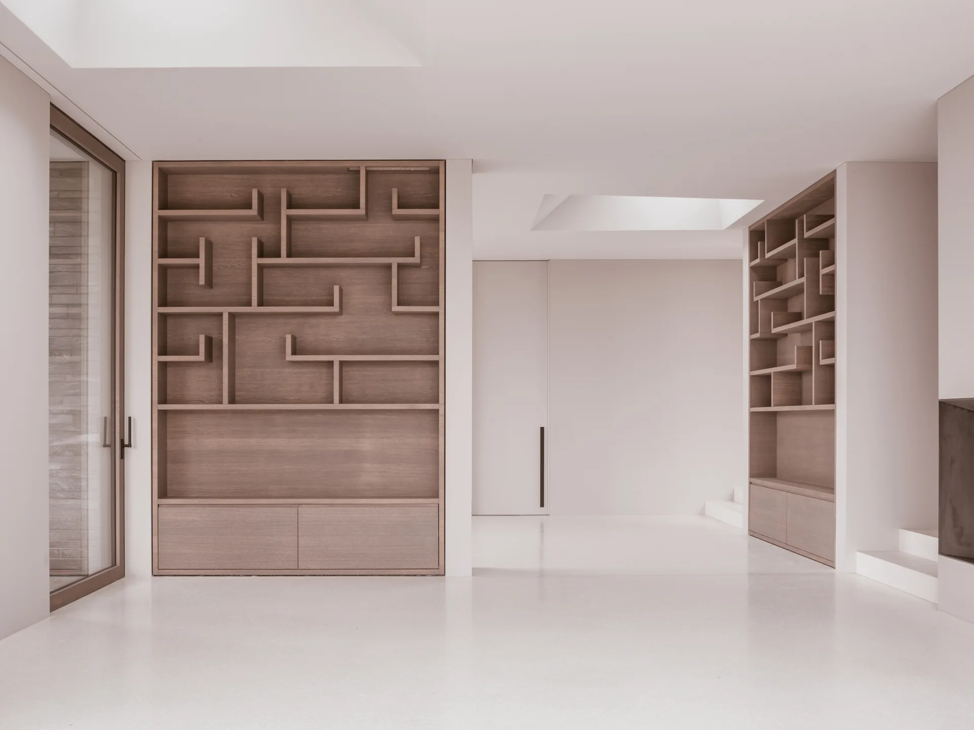 Two custom light wood bookshelves with a geometric divider design are built into matte white walls under a recessed ceiling skylight.