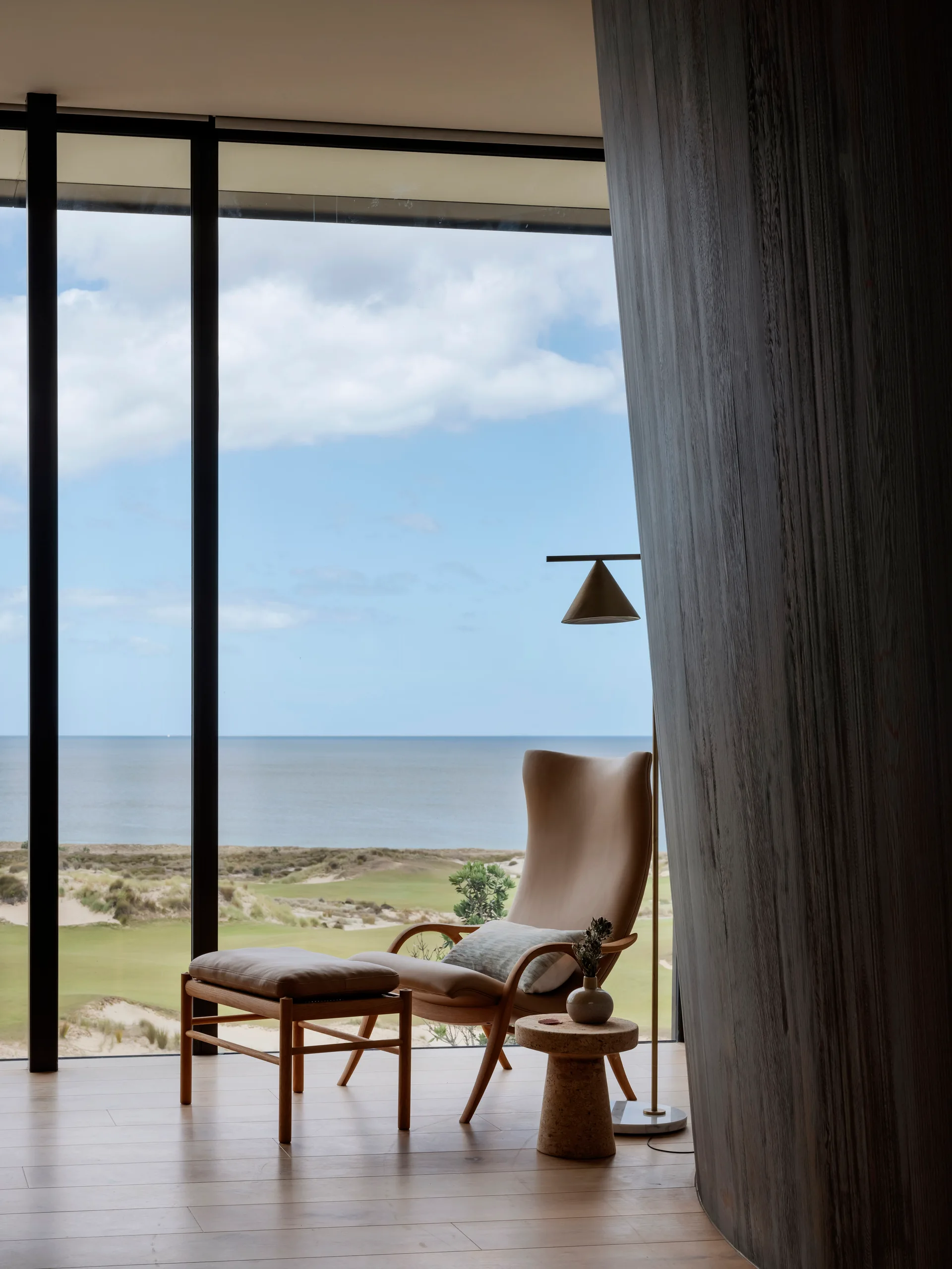 A tan leather armchair and ottoman sit next to a curved wood partition wall in front of a floor-to-ceiling window overlooking the ocean.