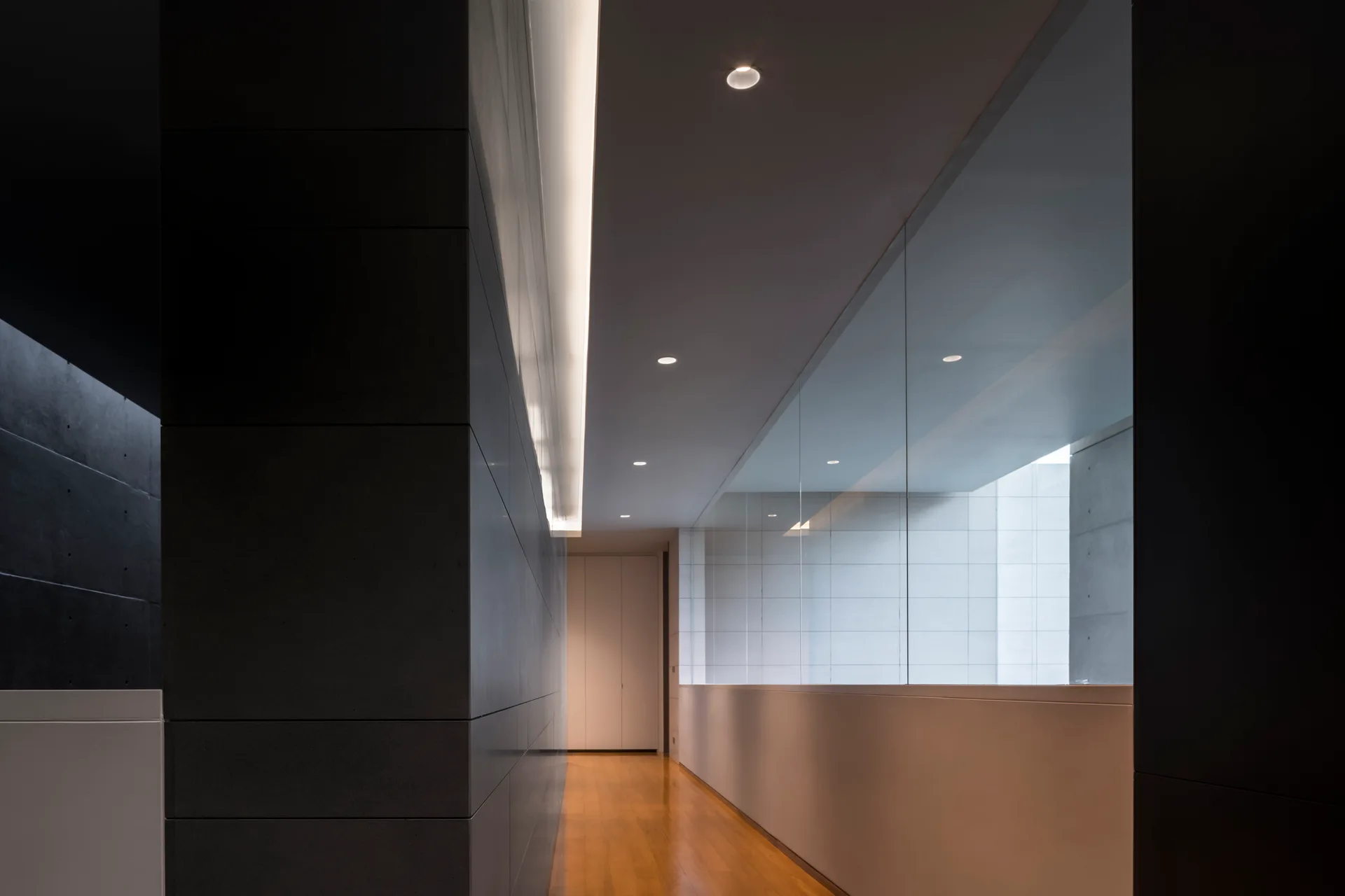 A minimalist hallway features charcoal grey wall panels, polished wood flooring, a glass partition, and warm recessed architectural lighting.