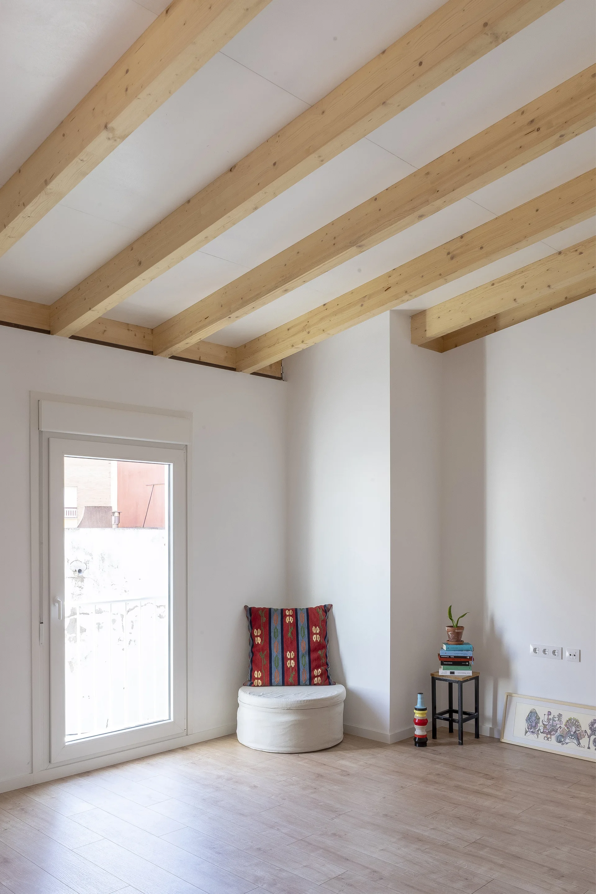 A minimalist white room features exposed light wood ceiling beams, a round white stool with a patterned red pillow, and a small side table.