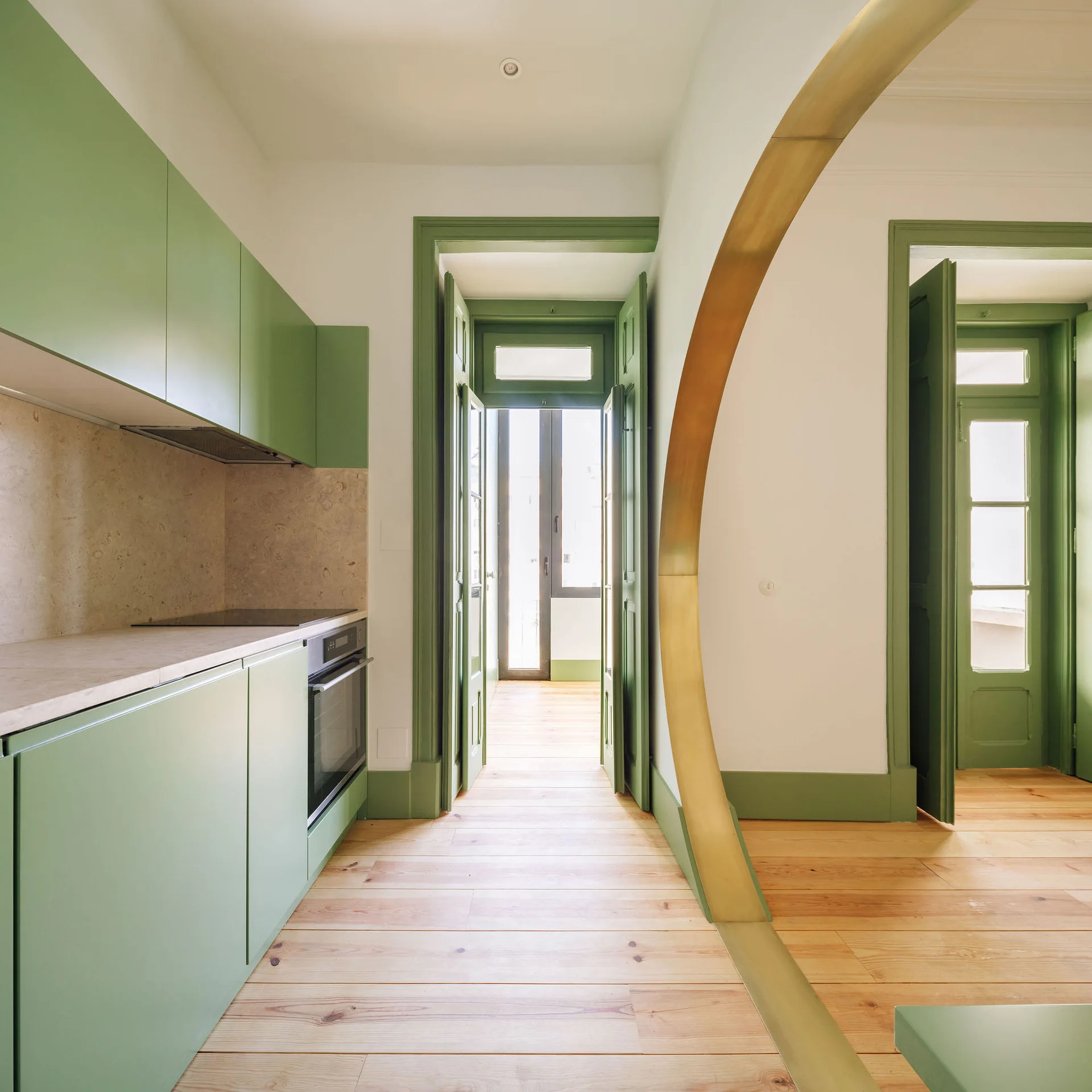 A modern kitchen with sage green cabinets, light wood flooring, green-trimmed doorways, and a large curved brass archway.