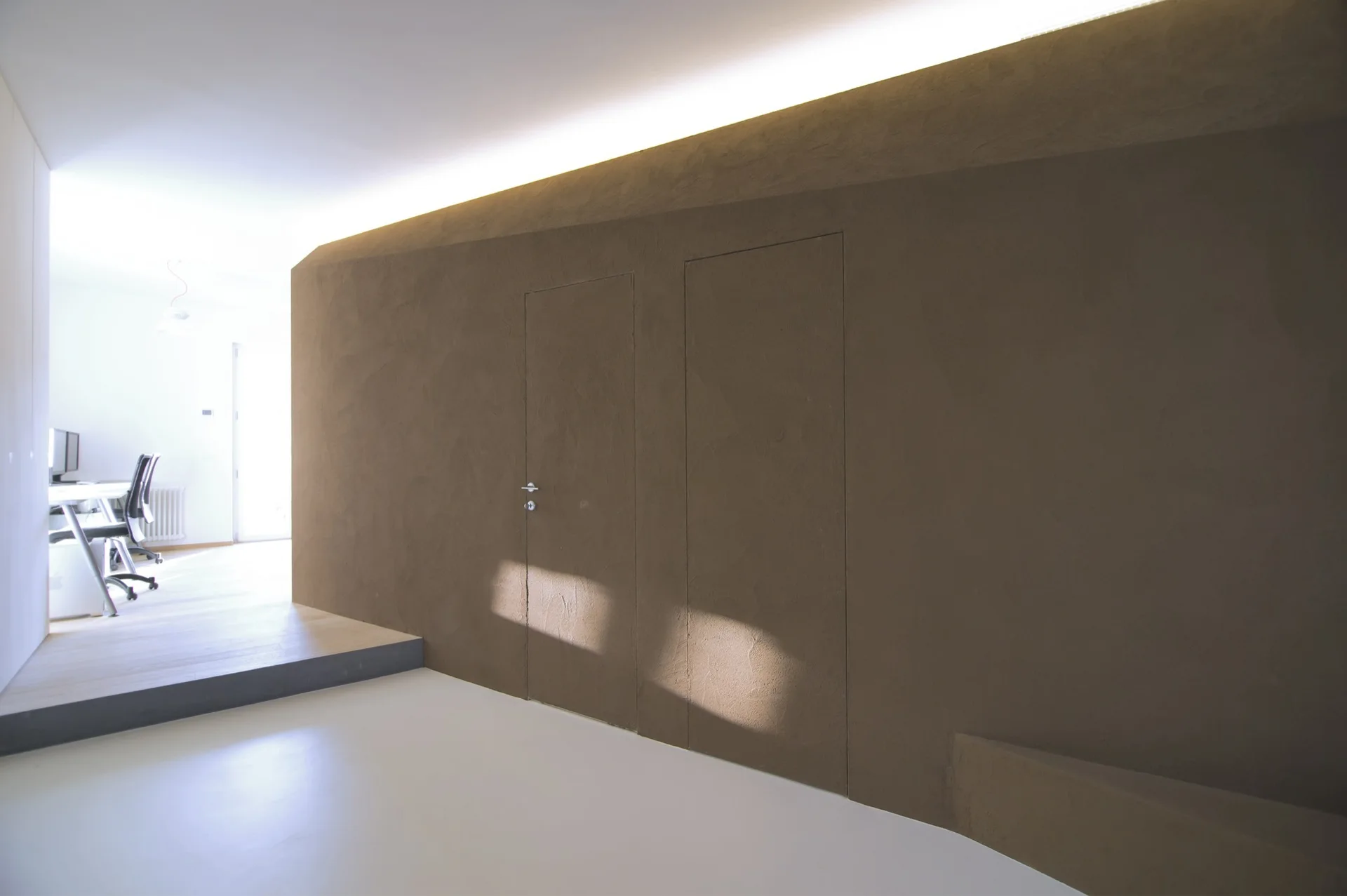A minimalist hallway features a textured brown wall with two flush-mounted concealed doors beneath bright cove lighting.