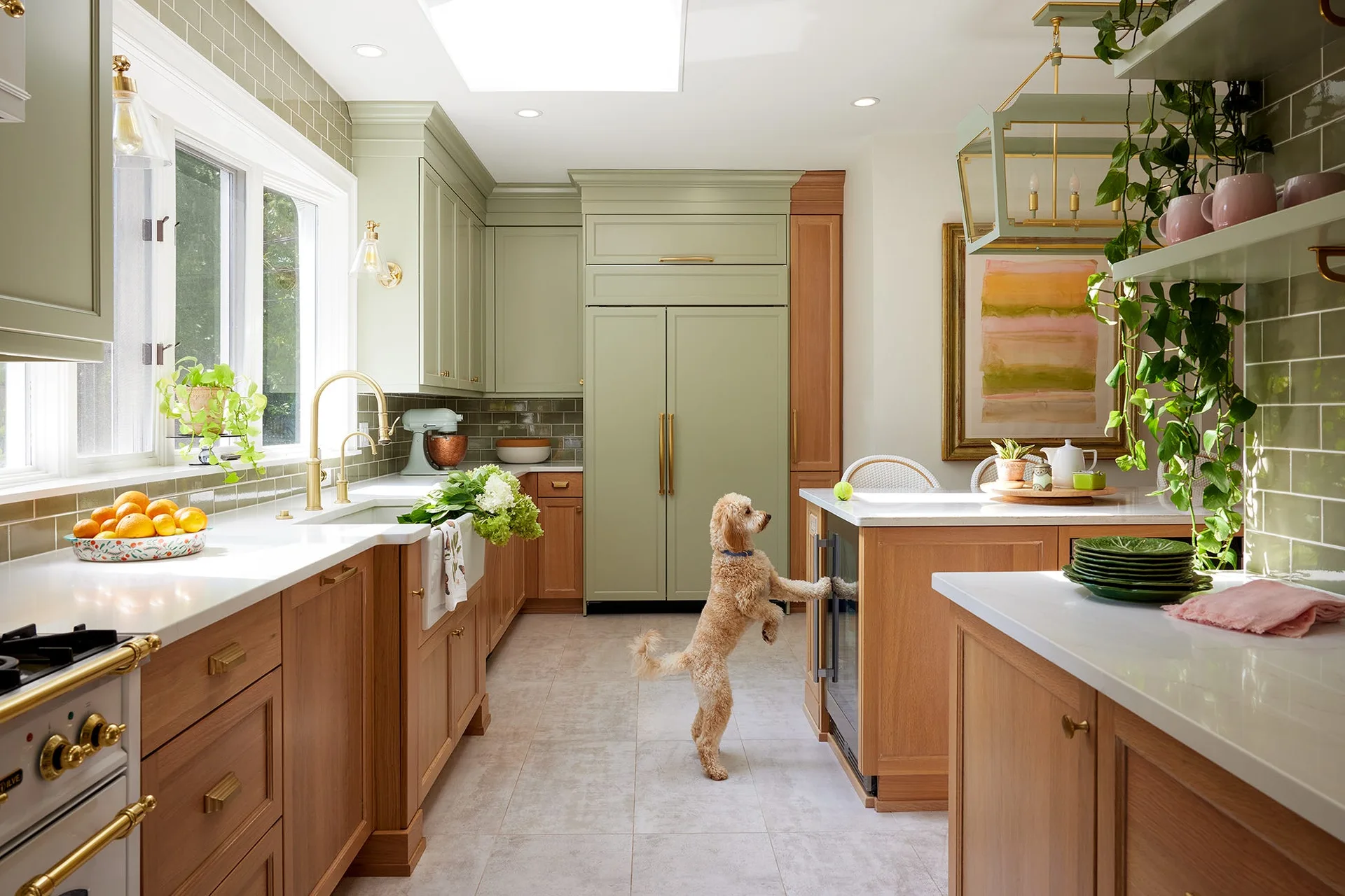 A bright kitchen features sage green upper cabinets, natural wood lower cabinets, green subway tile backsplash, and a dog standing near a wooden island.