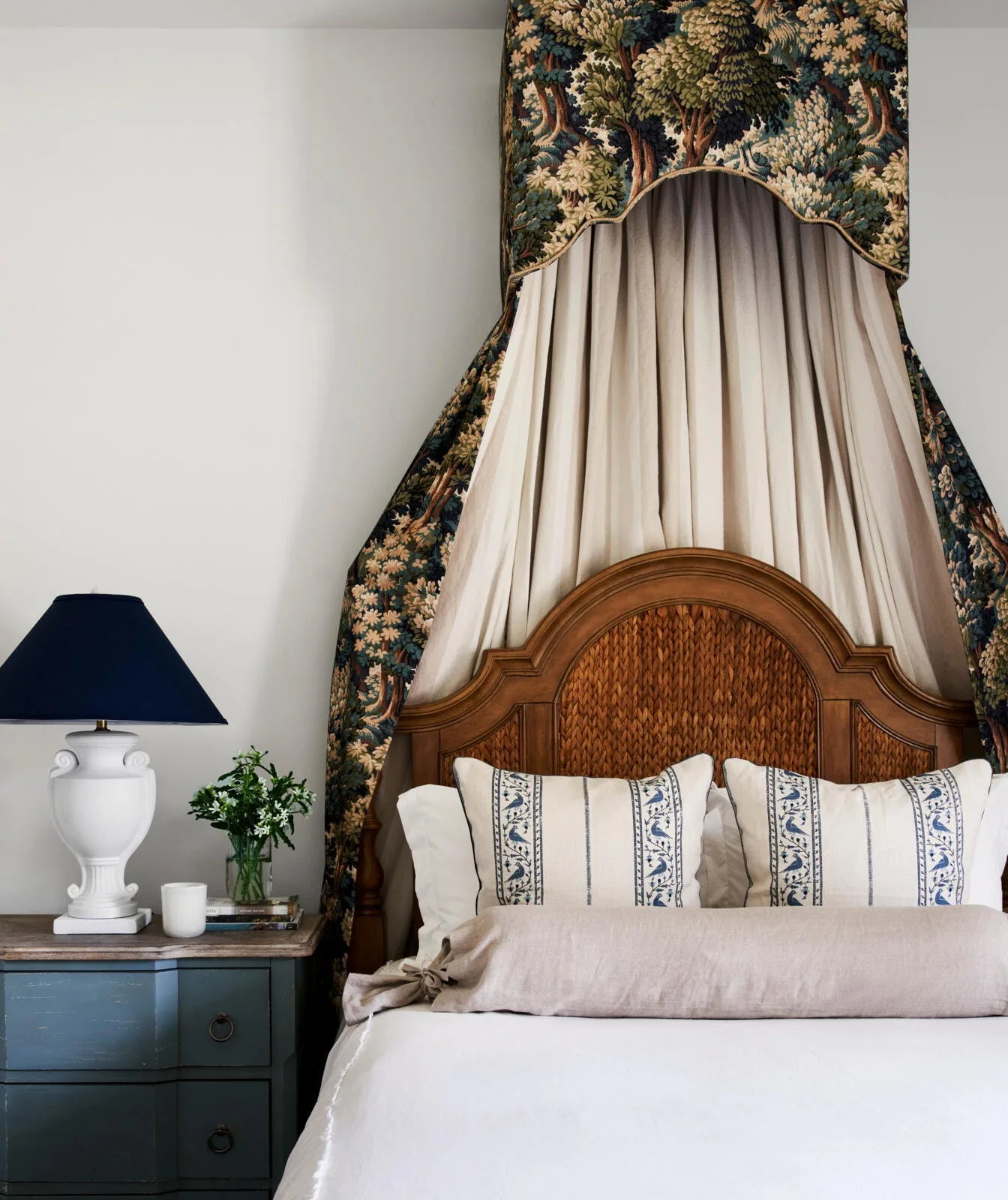 A traditional wooden bed features a woven headboard and a patterned tapestry canopy next to a blue nightstand with a white table lamp.