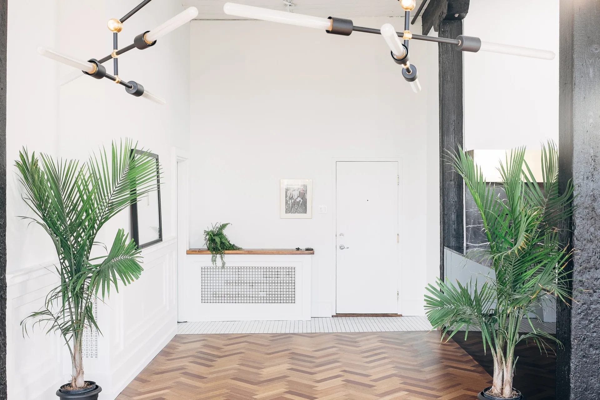 A bright entryway features brown herringbone wood flooring, white paneled walls, a modern linear chandelier, and two large potted palms.