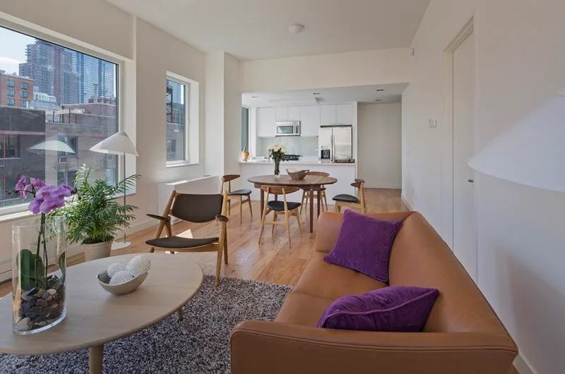 An open-concept living area features a tan leather sofa with purple pillows, a light wood coffee table, and a round wooden dining table in the background.