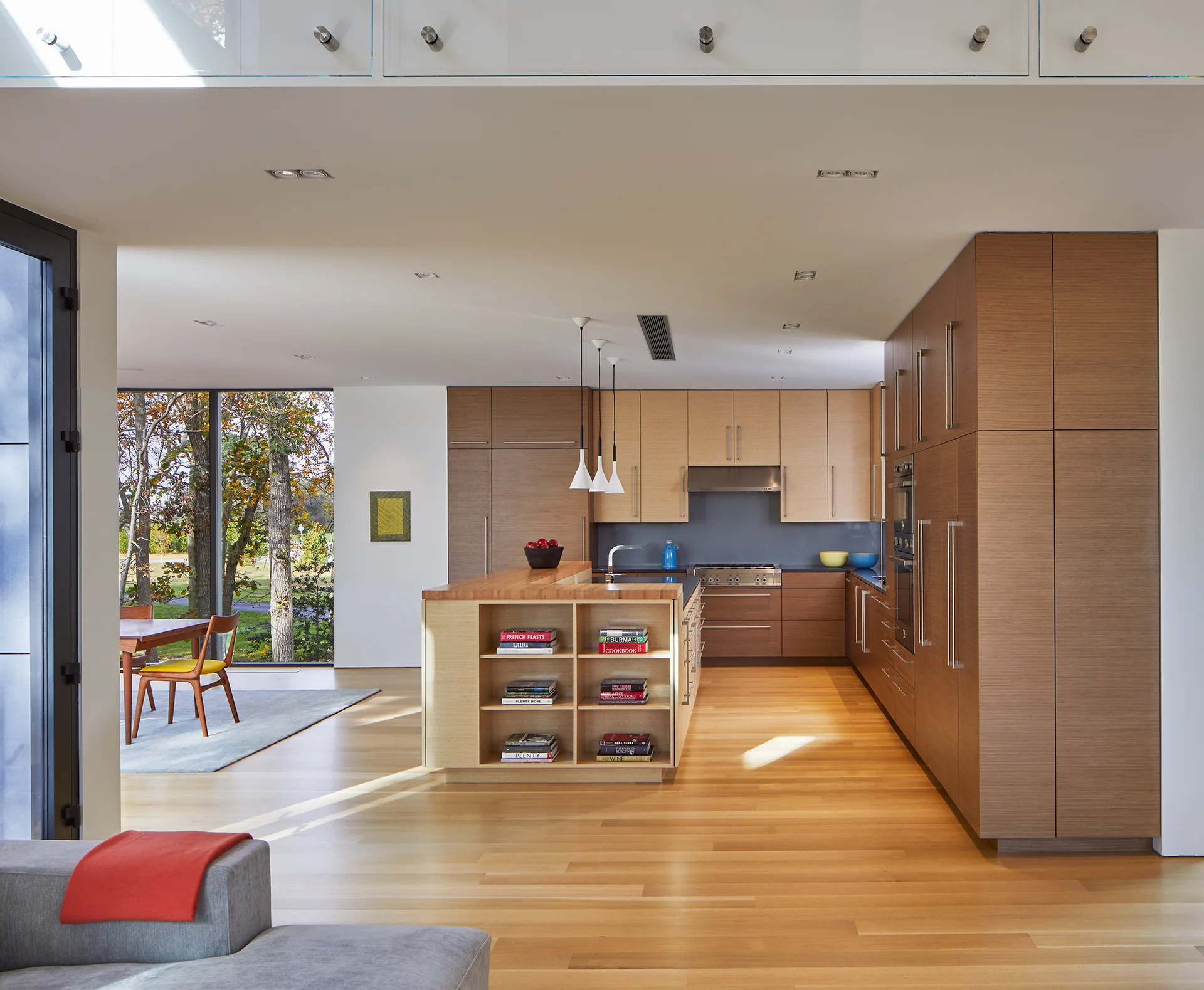 A modern open-concept kitchen features two-tone wood cabinetry, a central island with built-in bookshelves, and light hardwood floors.