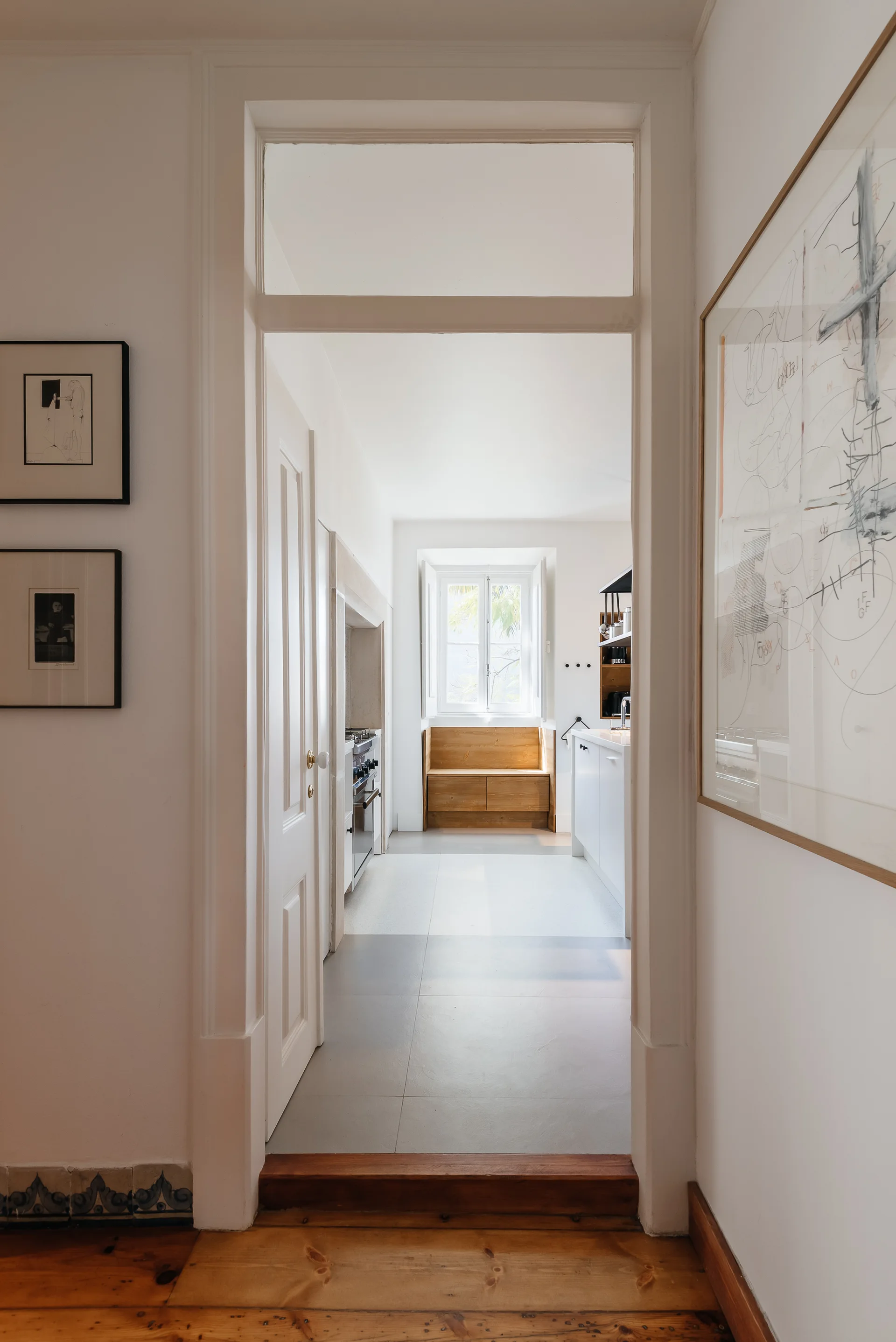 A view through a white doorway into a bright kitchen featuring white cabinets, grey floor tiles, and a built-in wooden bench under a window.