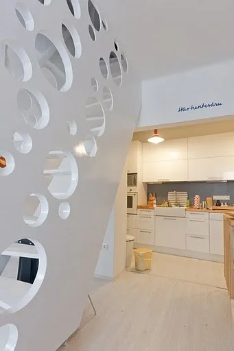 A modern white kitchen with light wood floors and white cabinets, seen behind a white staircase partition wall featuring several circular cutouts.
