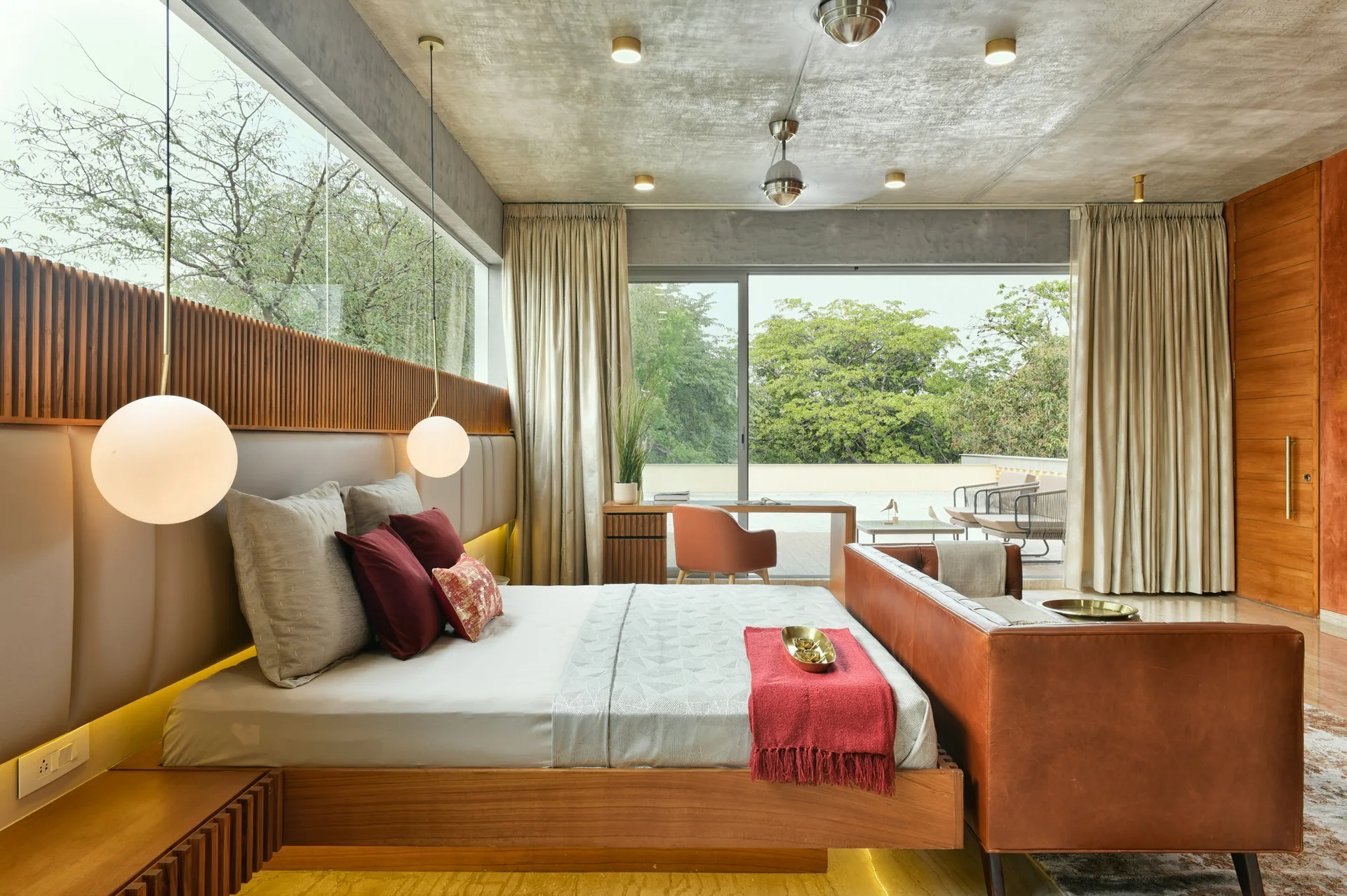 A contemporary bedroom features a wood platform bed, leather sofa, concrete ceiling, and large sliding glass doors opening to a terrace.