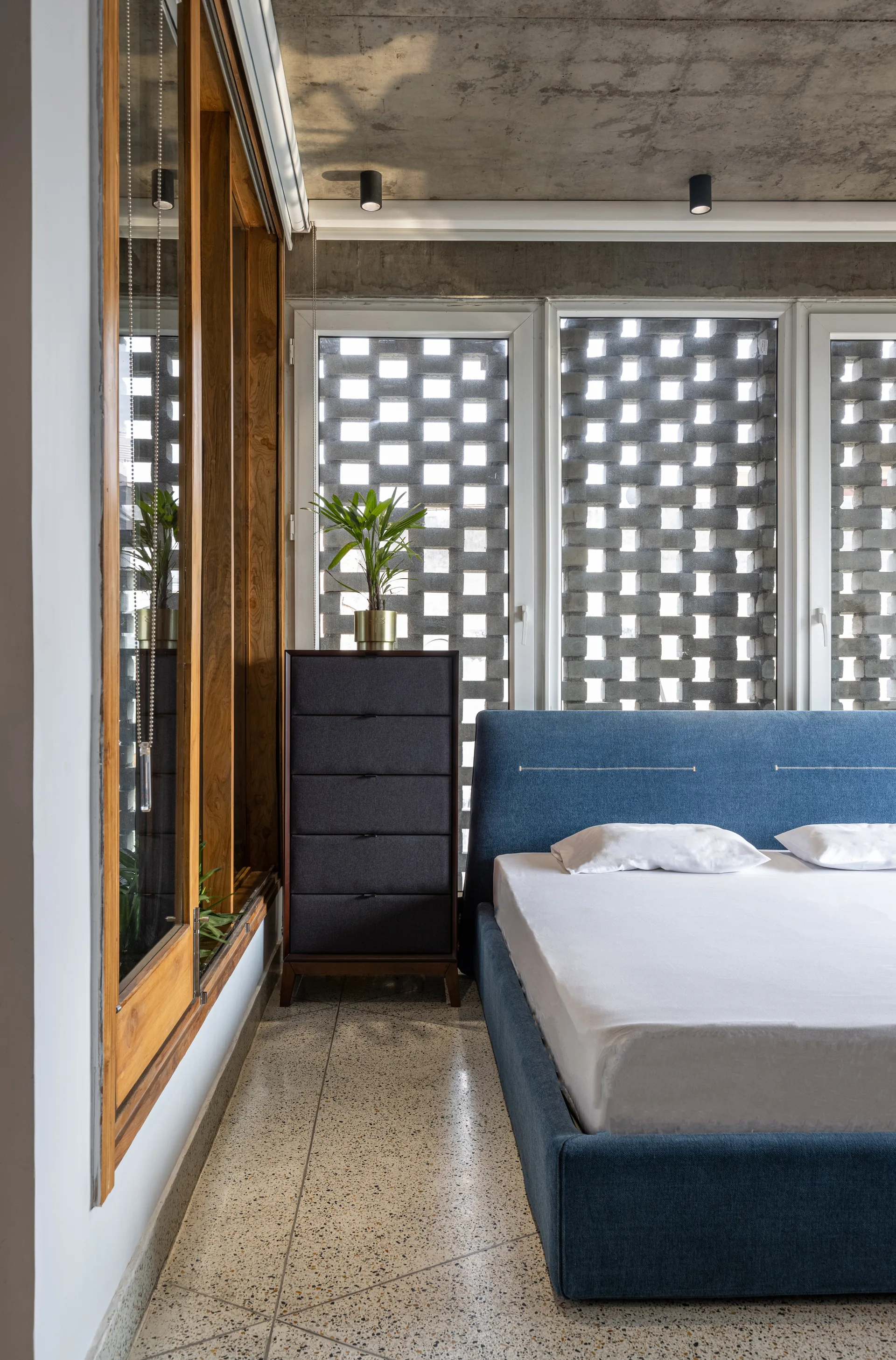 A modern bedroom with a blue upholstered bed, terrazzo floor, unpainted concrete ceiling, and a concrete breeze block window screen.