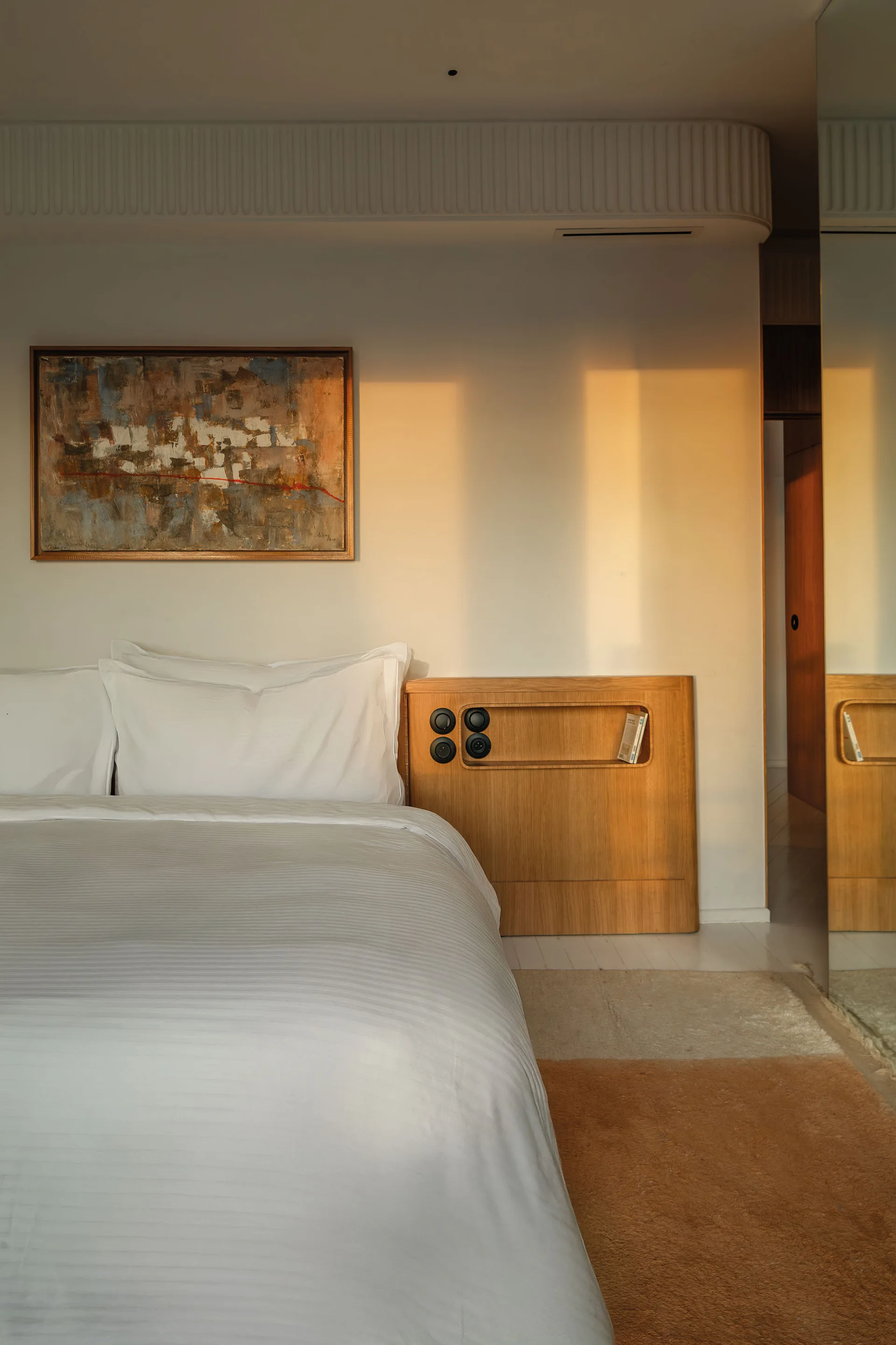 A minimalist bedroom features a bed with white bedding, an integrated wood headboard with a recessed shelf, and an abstract painting on the wall.