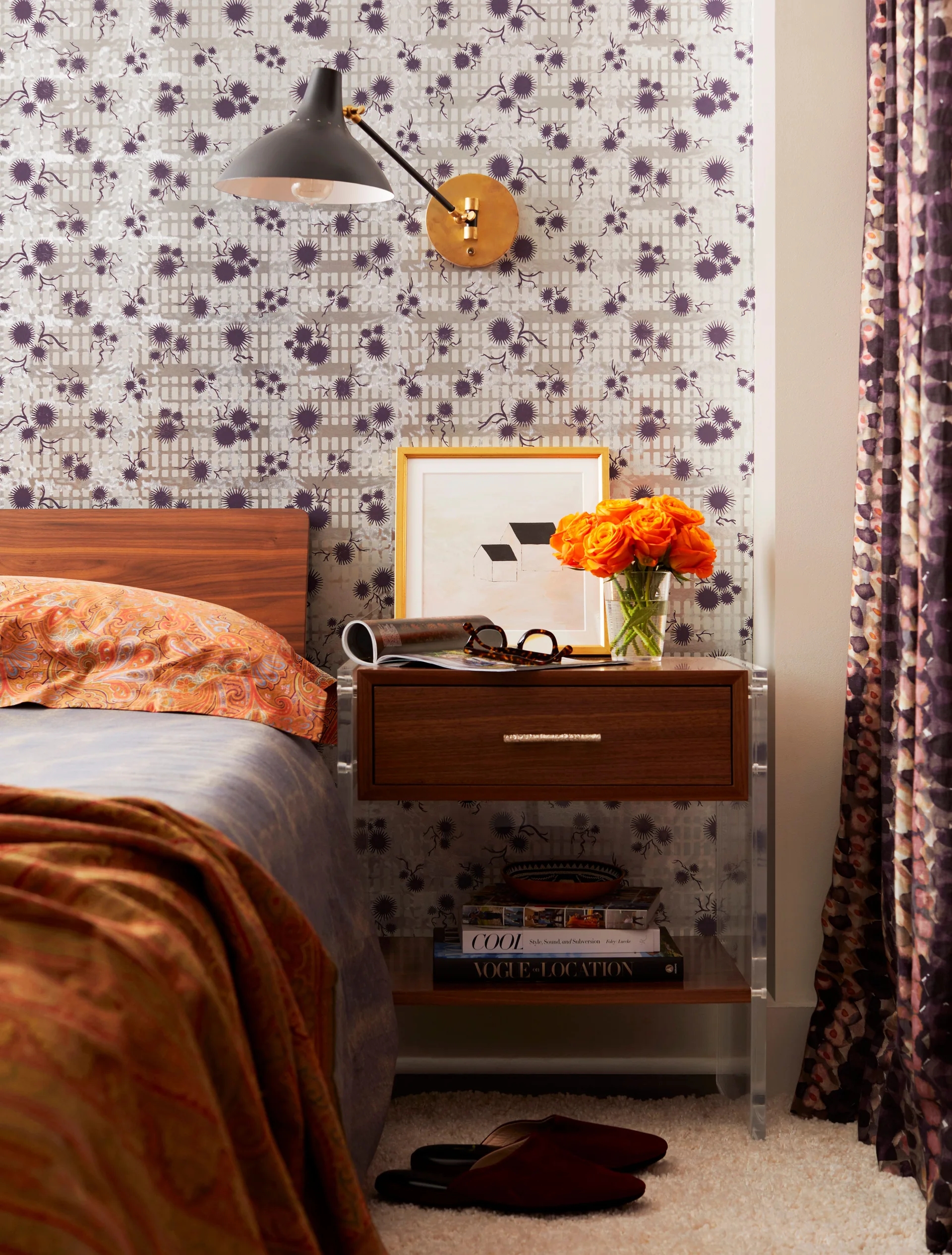 A wooden nightstand with clear acrylic sides features orange roses and a framed drawing against patterned wallpaper.