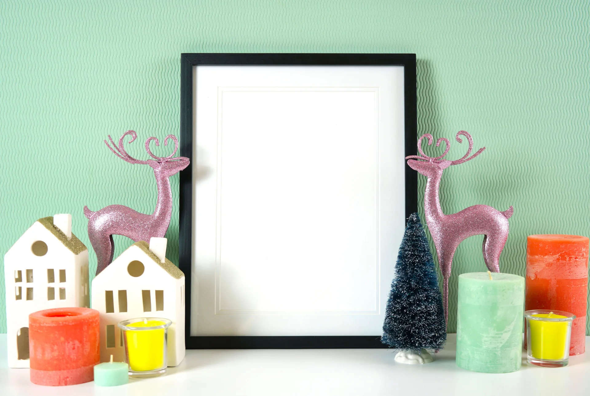 A black picture frame sits on a white tabletop surrounded by pink reindeer, toy houses, and colorful candles against a wavy mint green wall.