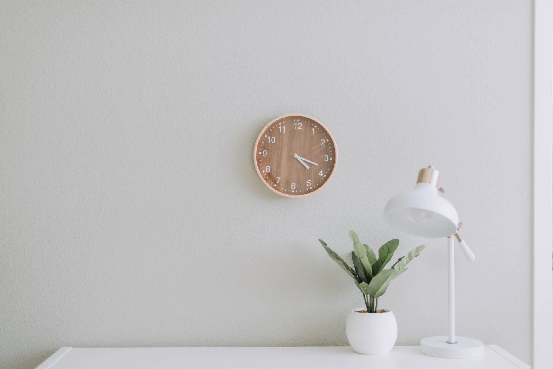A circular wooden clock hangs on a light grey wall above a white desk with a white task lamp and a small potted plant.