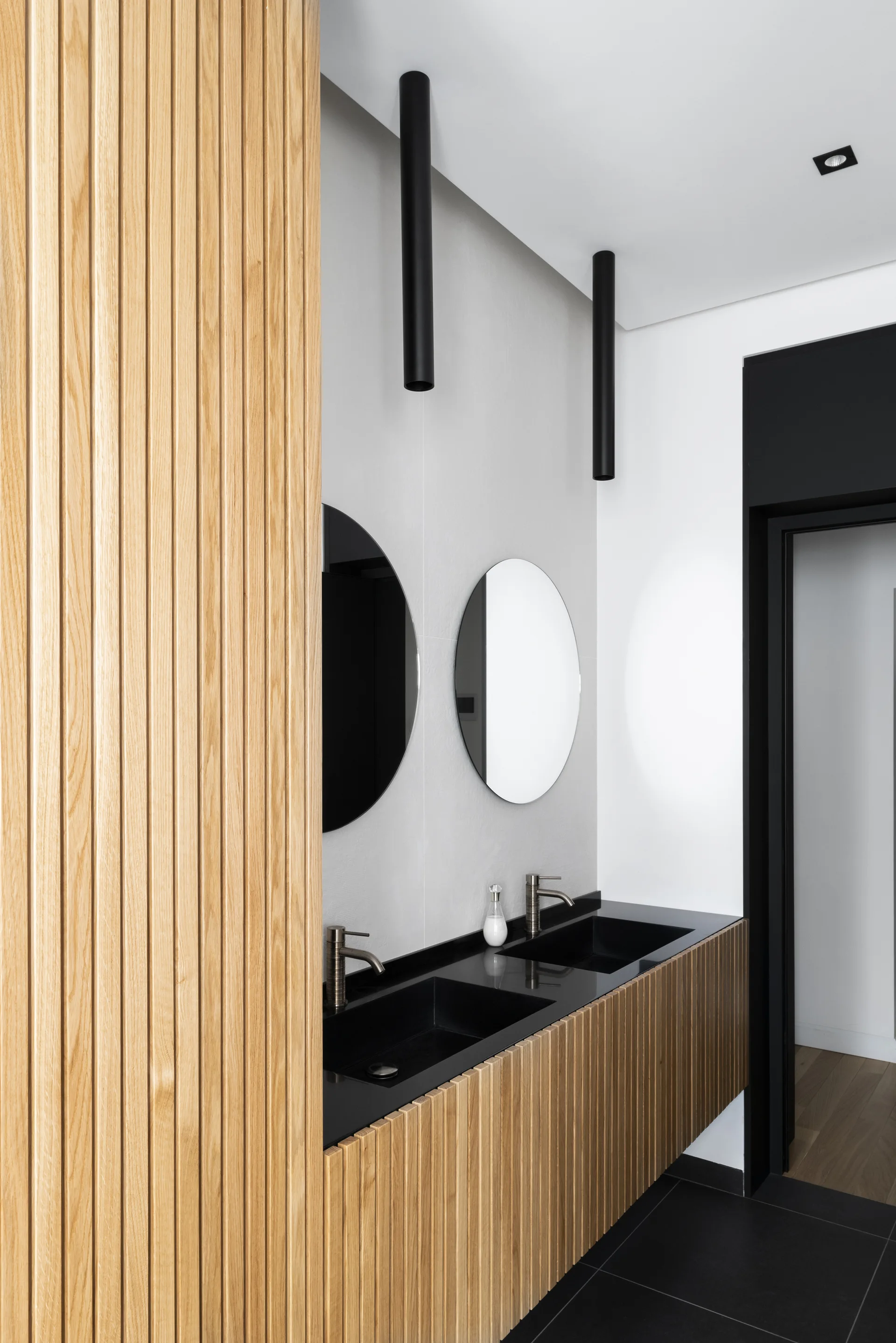 A modern bathroom features a double vanity with a light oak slatted facade, two black sinks, circular mirrors, and cylindrical black pendant lights.