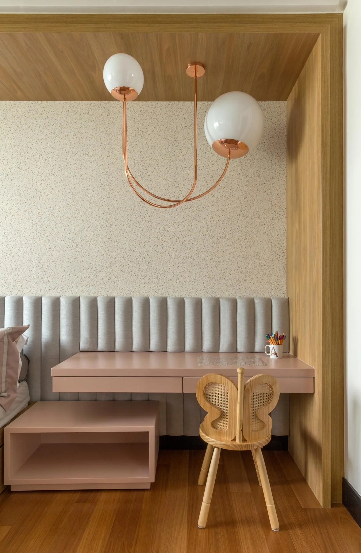 A children's study nook features a light pink built-in desk, a wooden chair with a butterfly back, and a channel-tufted headboard under a copper pendant.