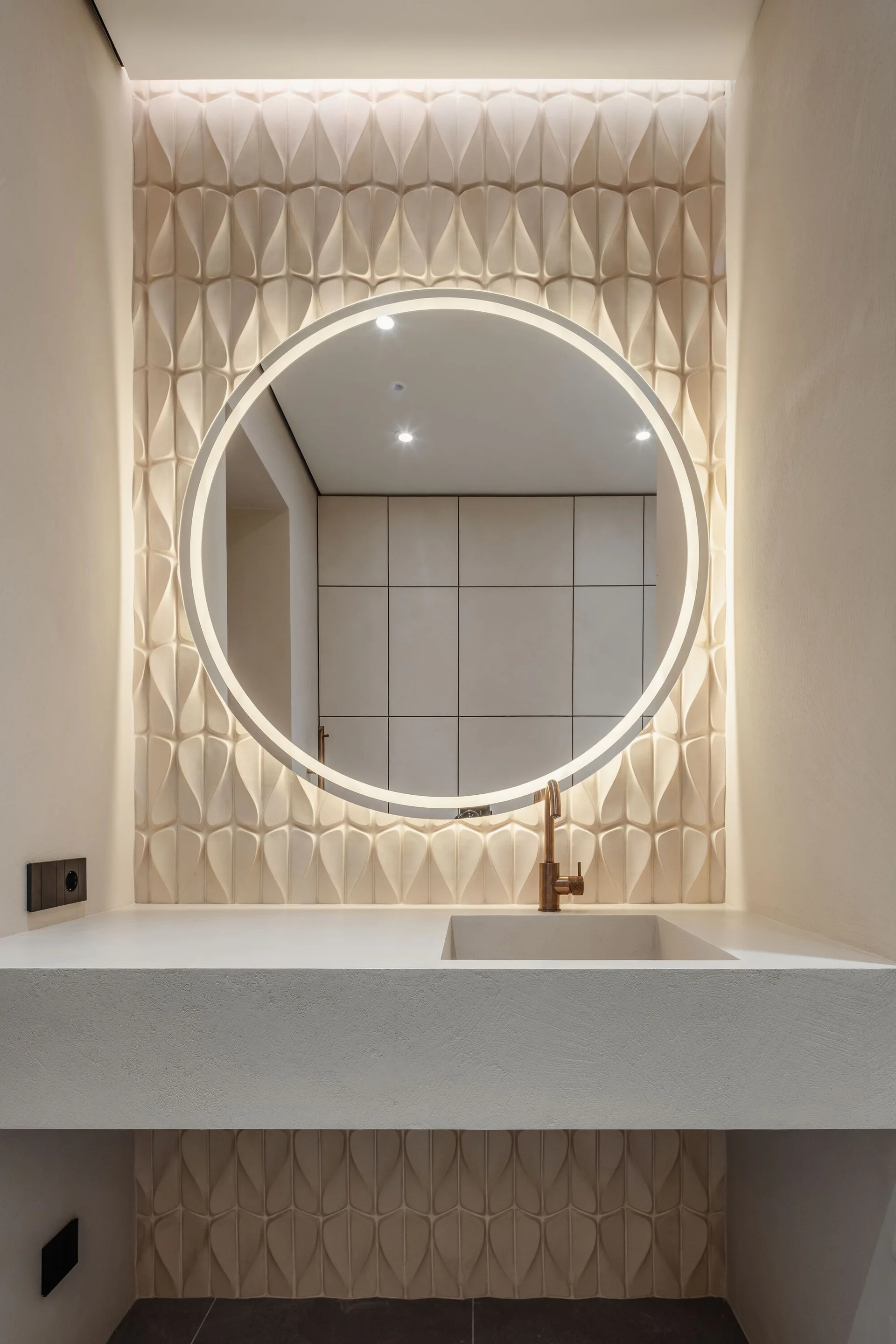 A floating cream bathroom vanity features an integrated sink, a bronze faucet, and a large backlit round mirror mounted on a textured geometric wall.