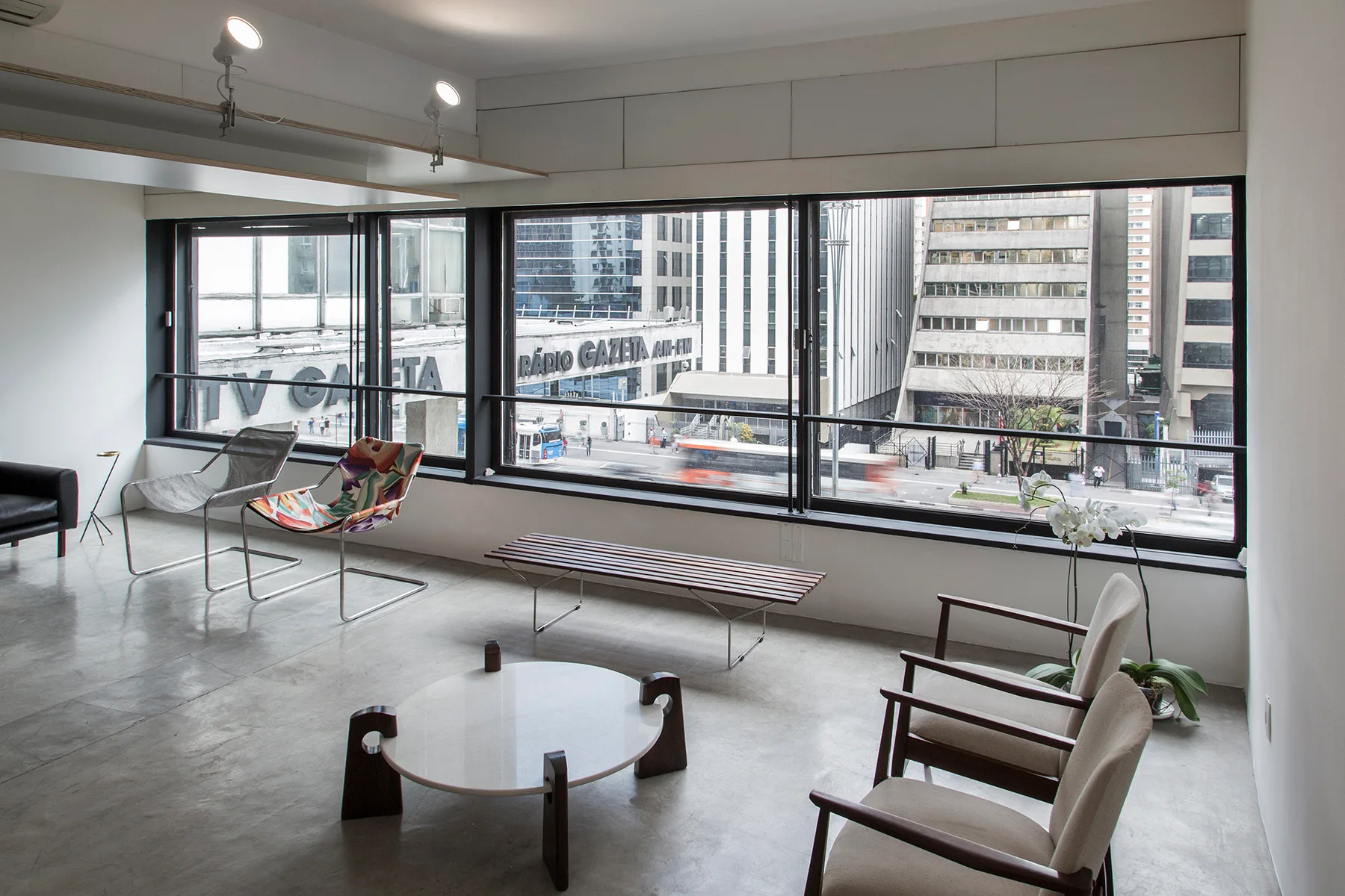 A modernist living area features a concrete floor, mid-century armchairs, a low round coffee table, and a wide window with views of a city street.