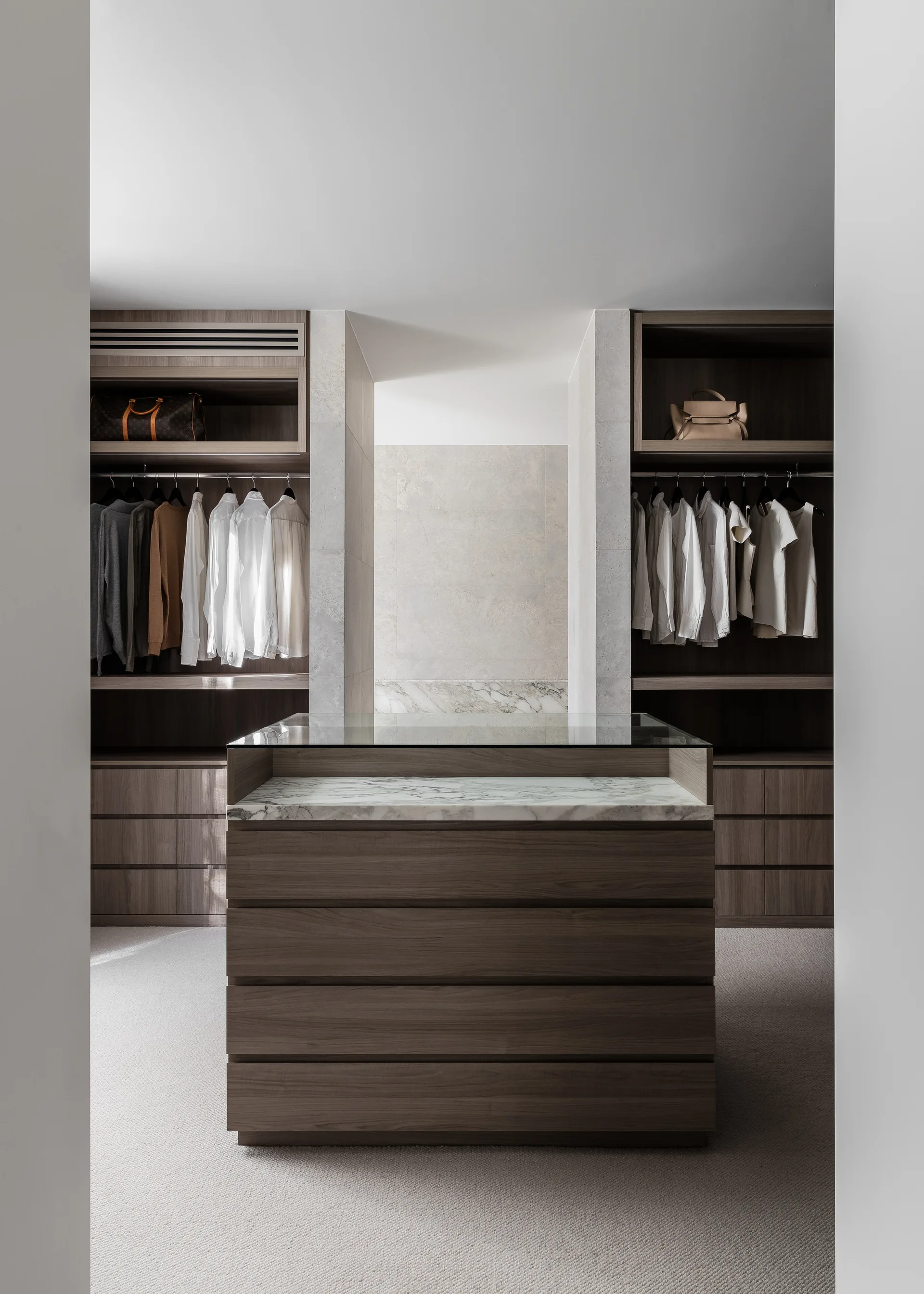 A symmetrical walk-in closet featuring built-in wood wardrobes on either side of a central drawer island with a veined marble top.